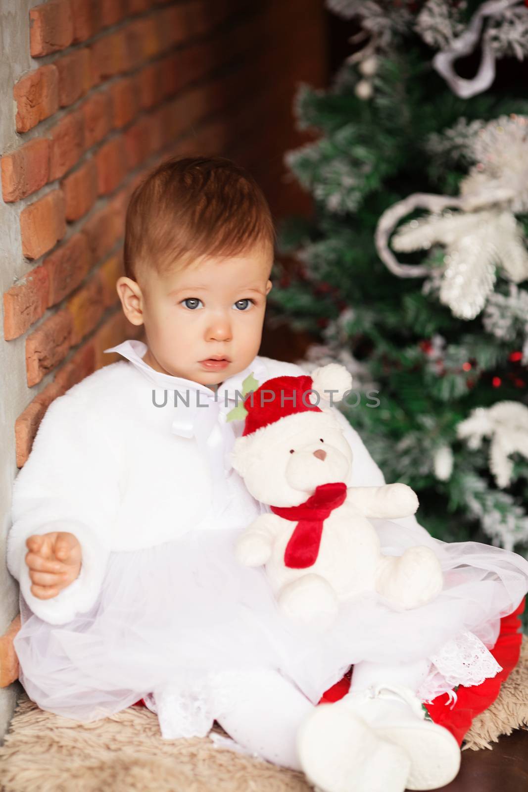Portrait of a beautiful baby girl with a soft teddy bear in Santa hat in the interior with Christmas decorations.