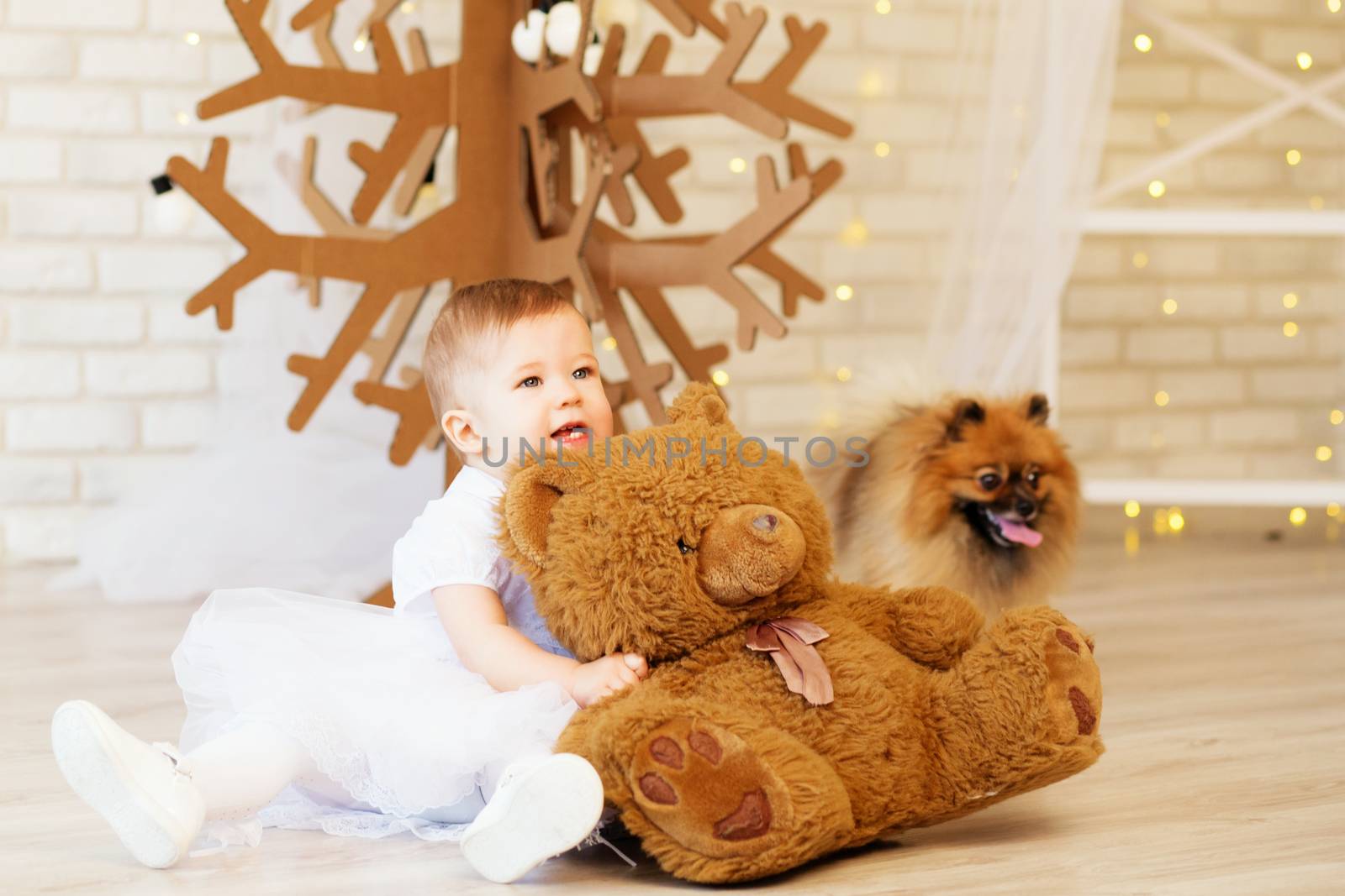 Portrait of a beautiful baby girl with a soft brown teddy bear in the interior with Christmas decorations. Kid and puppy
