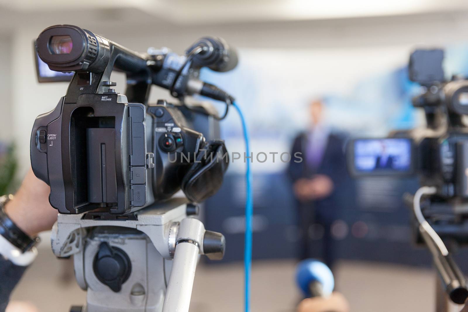 Video camera in focus, blurred spokesperson in background. Corporate news conference.