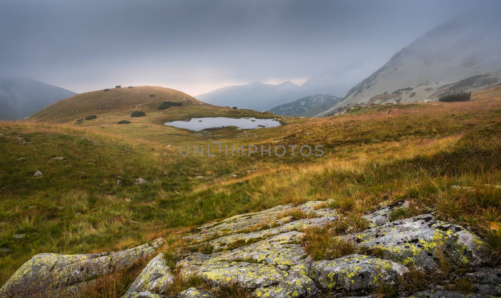 Foggy Mountain Landscape with a Tarn and a Rock in Foreground at Sunset