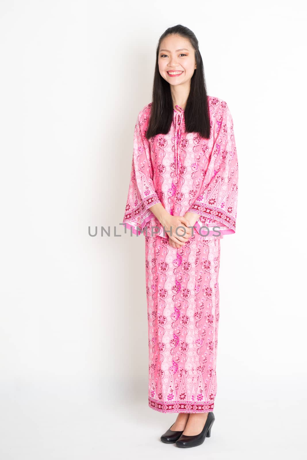 Portrait of young southeast Asian girl in traditional Malay batik dress smiling, standing on plain background.