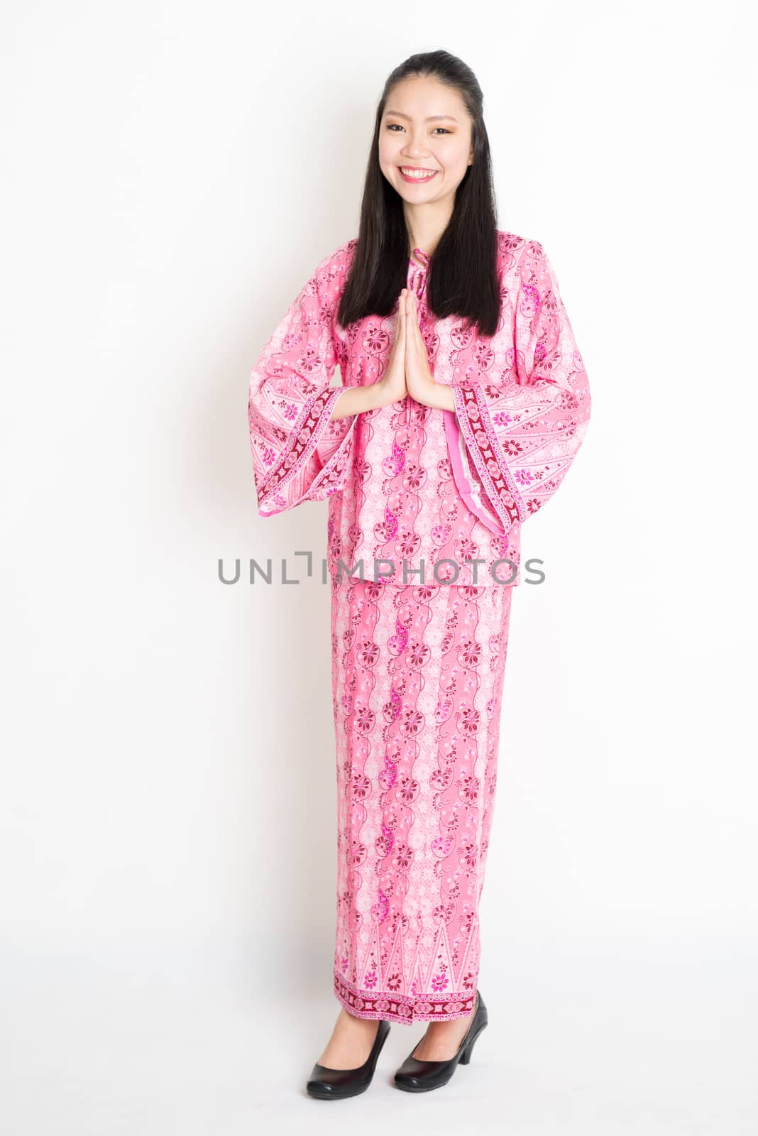 Portrait of young southeast Asian female in traditional Malay batik dress greeting, standing on plain background.