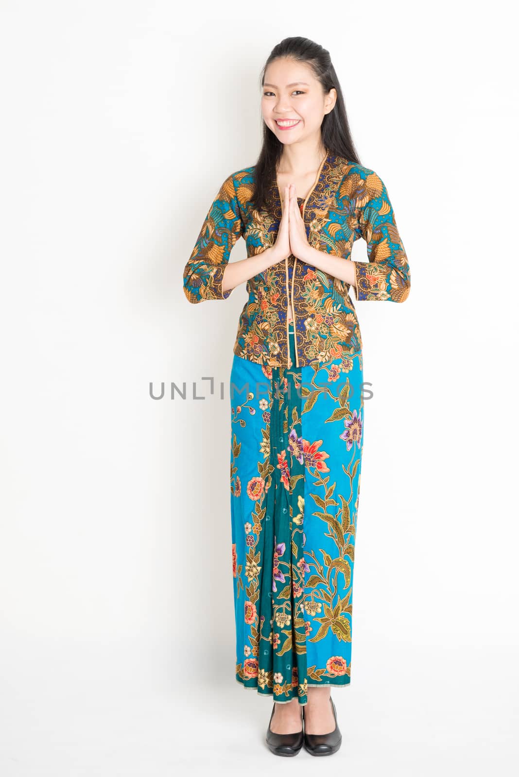 Portrait of young southeast Asian woman in traditional Malay batik kebaya dress greeting, full length standing on plain background.