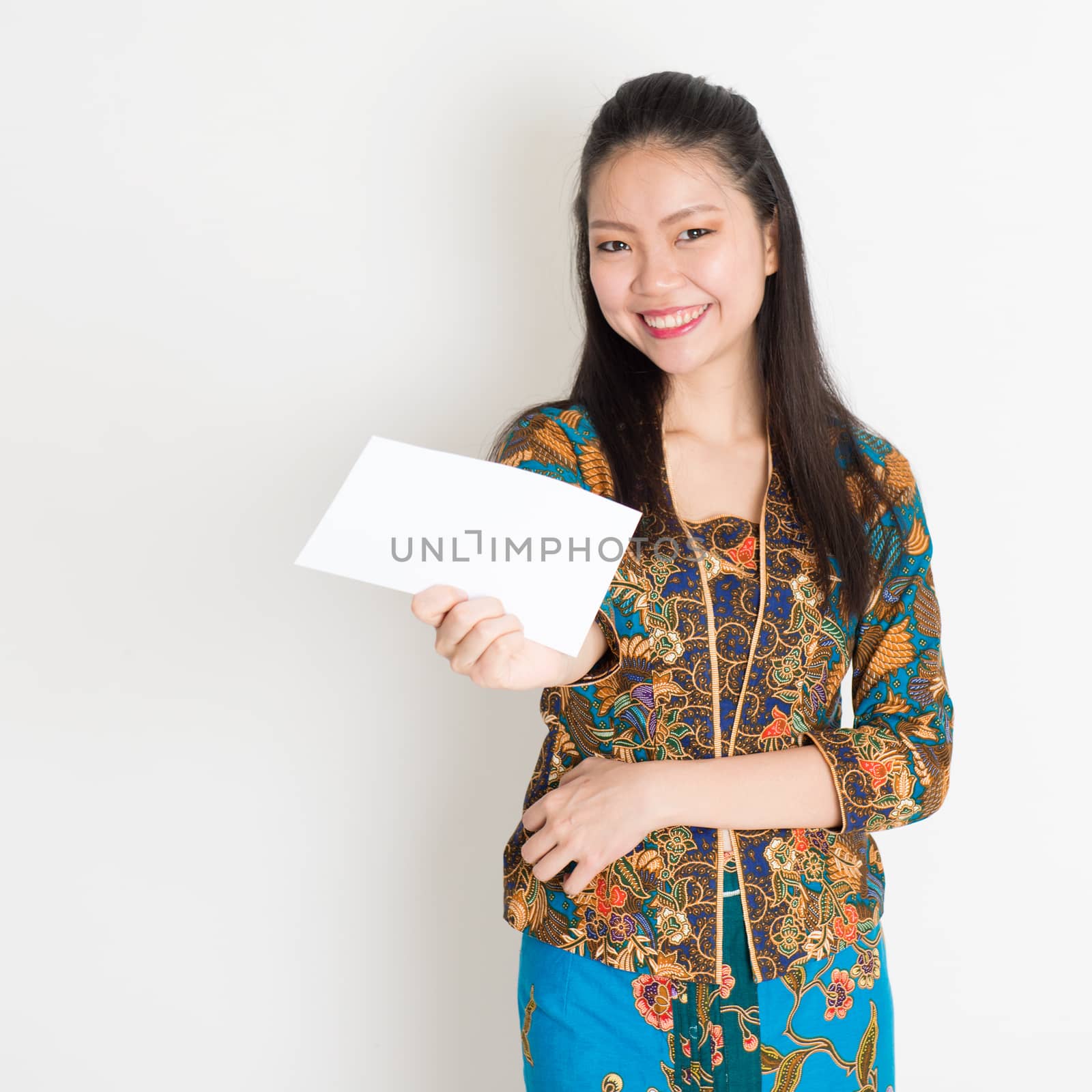 Portrait of young southeast Asian woman in traditional Malay batik kebaya dress hand holding a white blank paper card, standing on plain background.