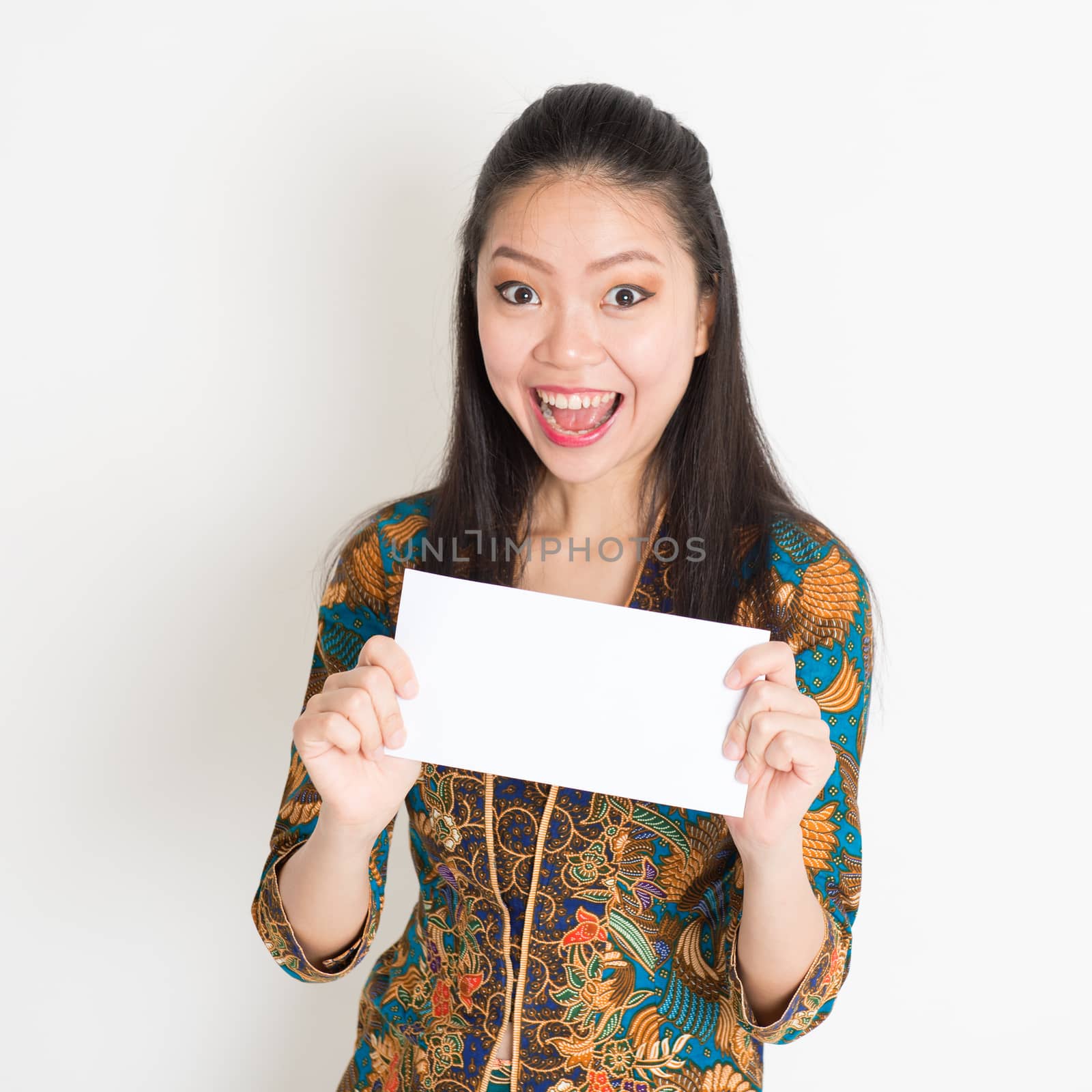 Portrait of young southeast Asian woman in traditional Malay batik kebaya dress hand holding a white blank paper card with surprised face expression, standing on plain background.