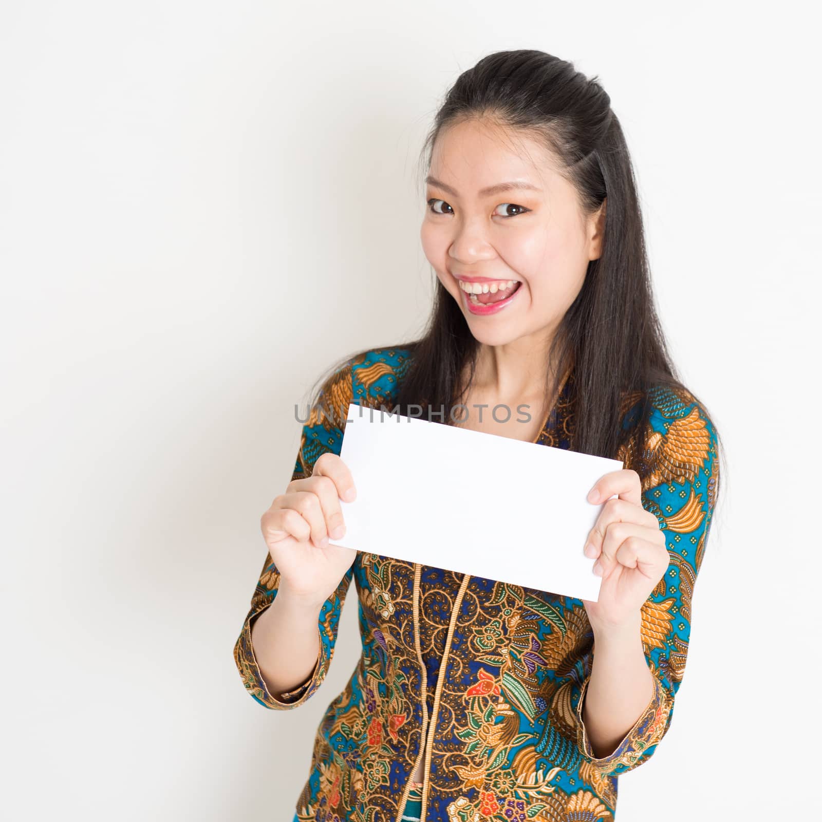Portrait of young southeast Asian girl in traditional Malay batik kebaya dress hand holding a white blank paper card with surprised face expression, standing on plain background.