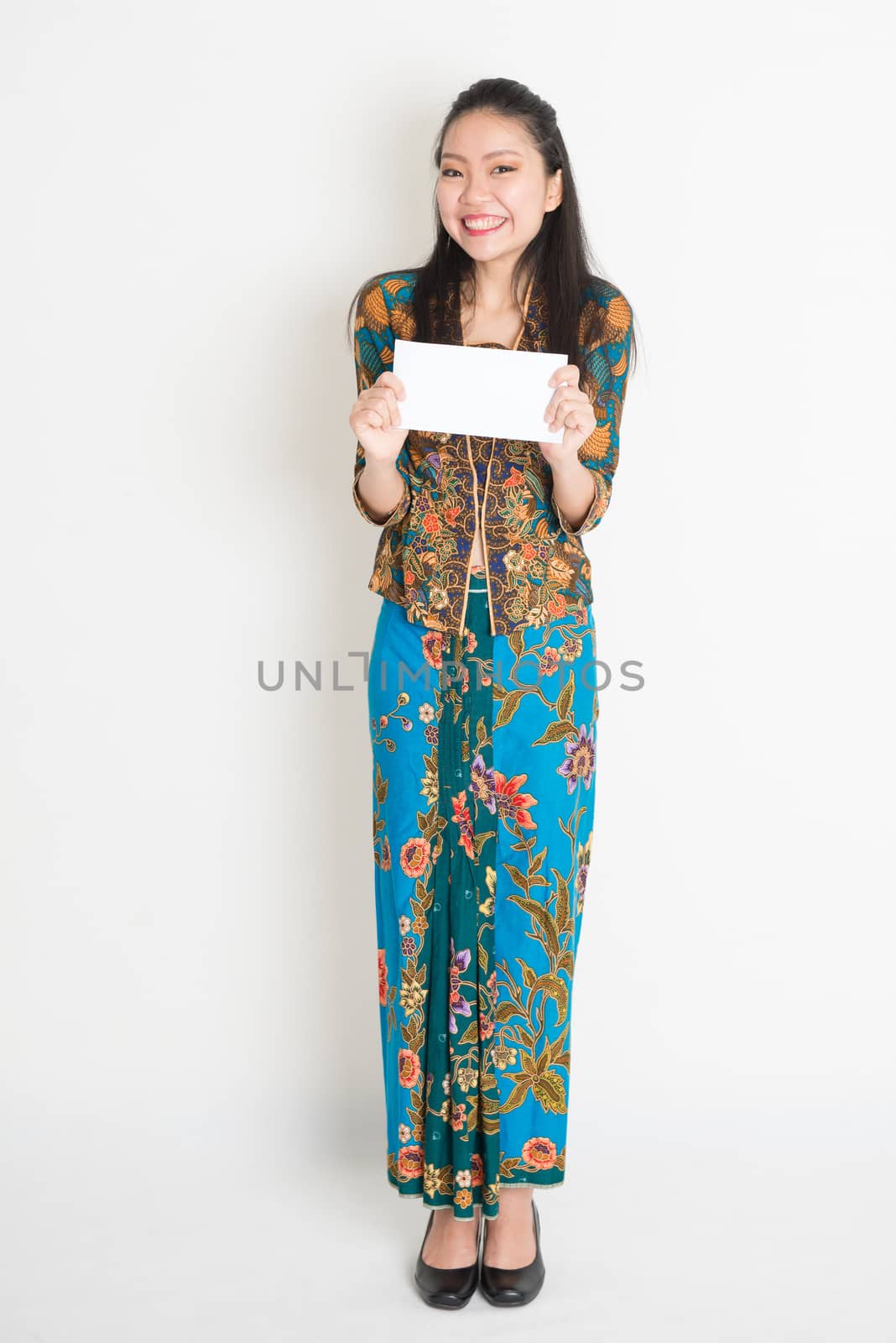 Portrait of young southeast Asian woman in traditional Malay batik kebaya dress hand holding a white blank paper card, full length standing on plain background.