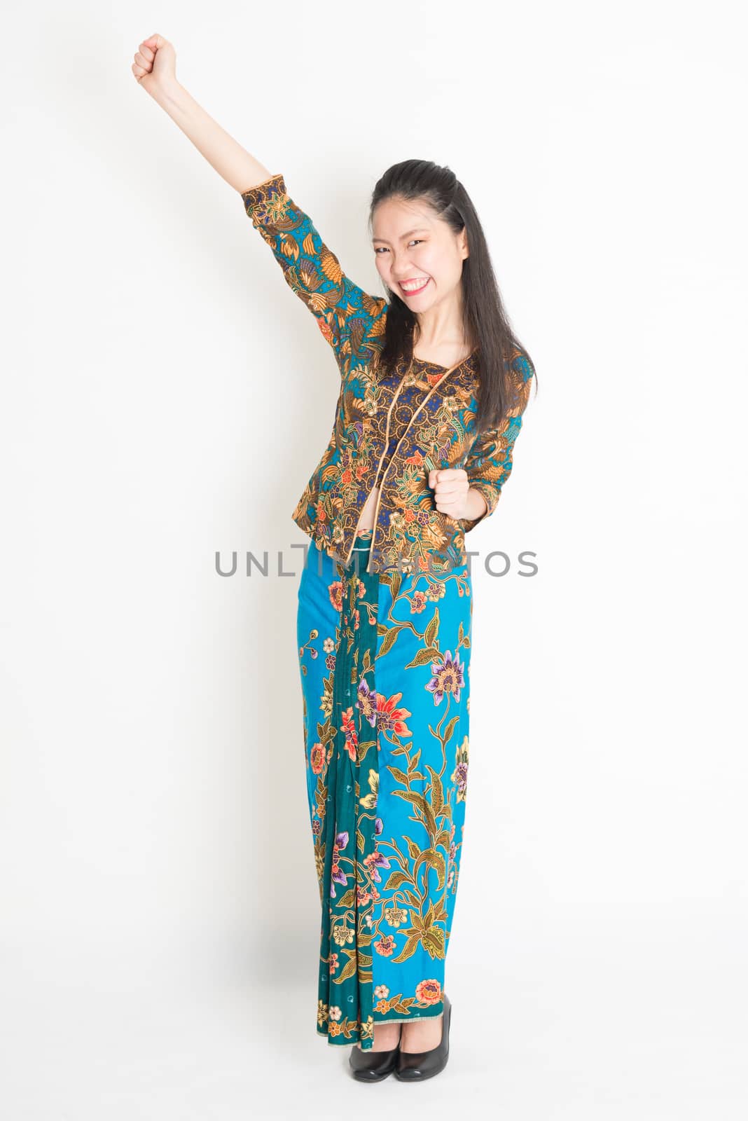 Portrait of young southeast Asian woman in traditional Malay batik kebaya dress arm raised celebrating success, full length standing on plain background.