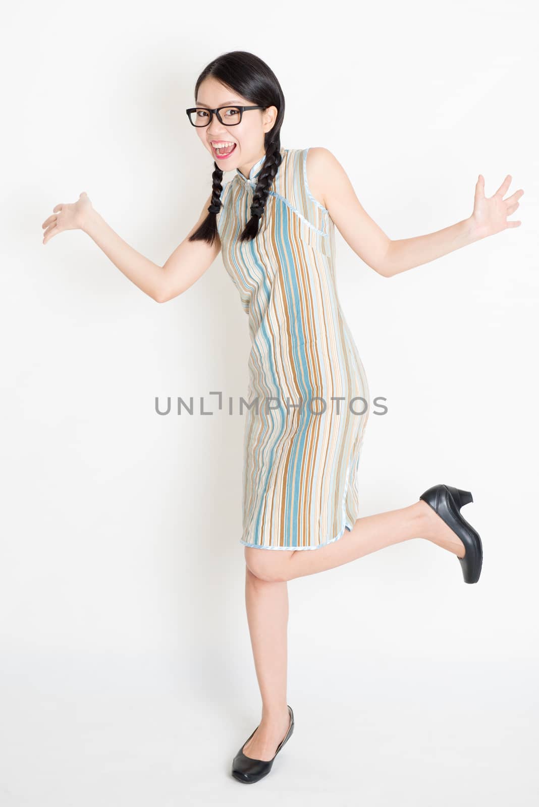 Portrait of excited young Asian girl in traditional qipao dress jumping around, full length standing on plain background.