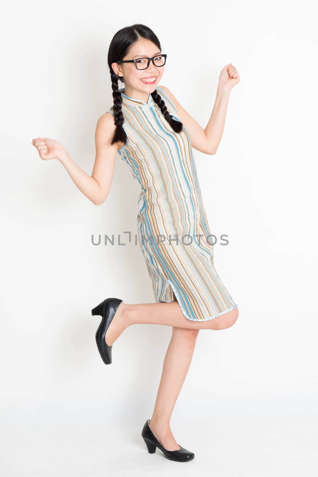 Portrait of excited young Asian girl in traditional qipao dress jumping around and hand holding something, full length standing on plain background.