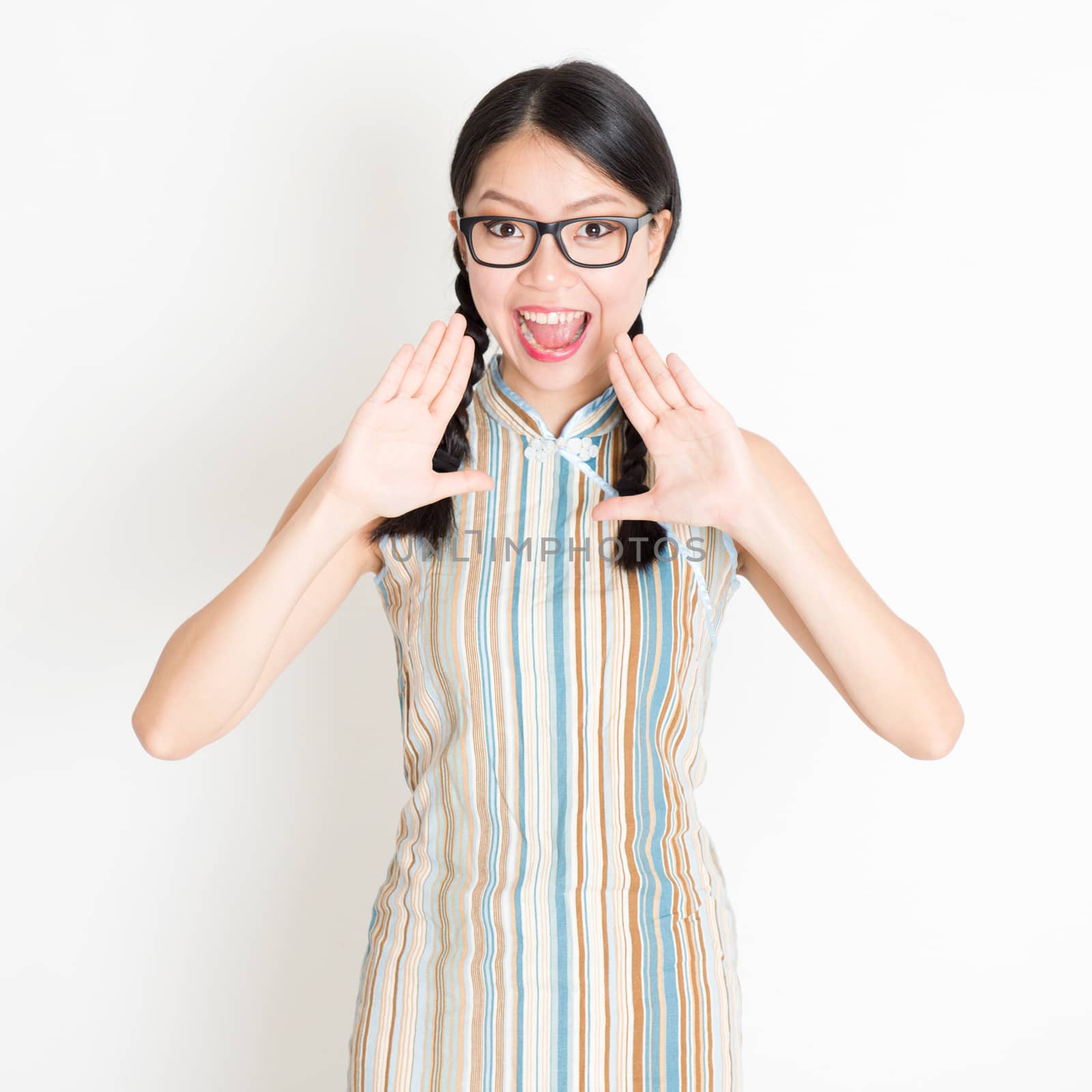 Portrait of young Asian girl in traditional qipao dress shouting, mouth opened wide, celebrating Chinese Lunar New Year or spring festival, standing on plain background.