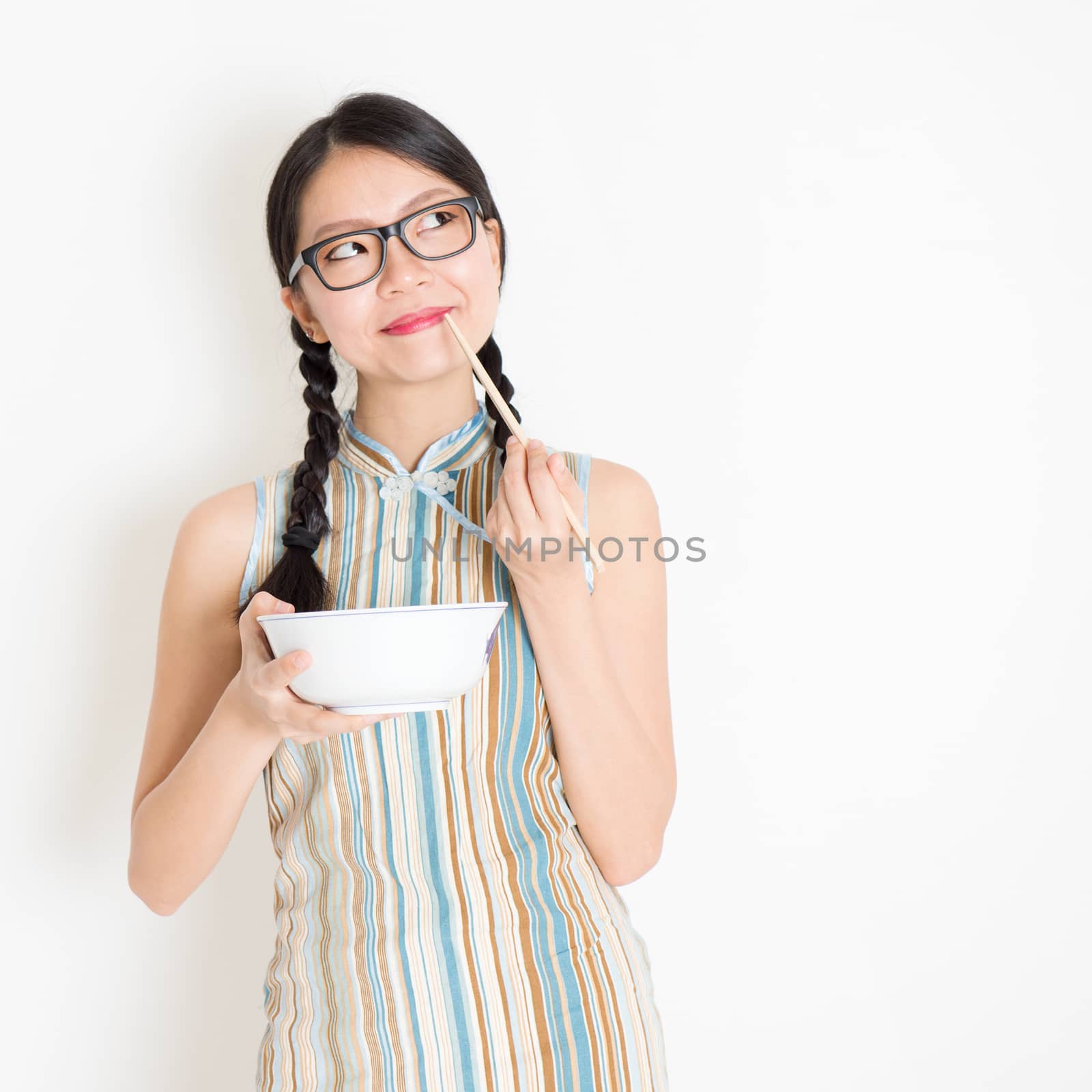 Portrait of young Asian woman in traditional qipao dress eating with chopsticks, celebrating Chinese Lunar New Year or spring festival, standing on plain background.