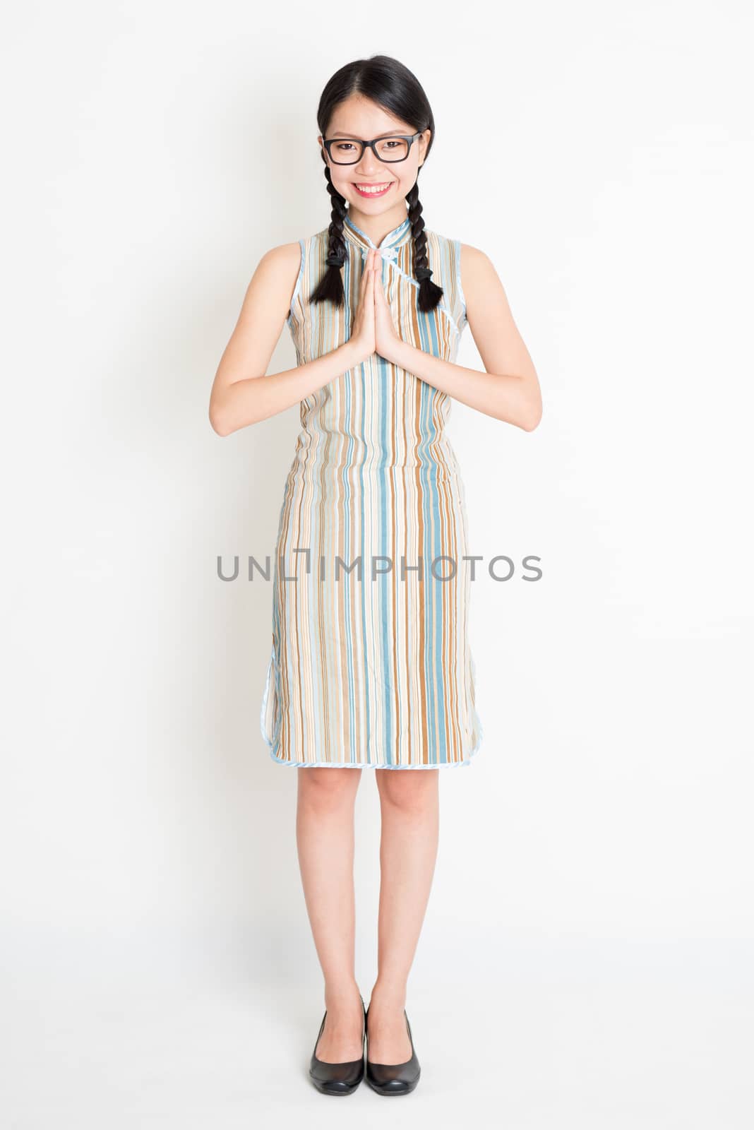 Portrait of young Asian girl in traditional qipao dress in greeting pose, full length standing on plain background.