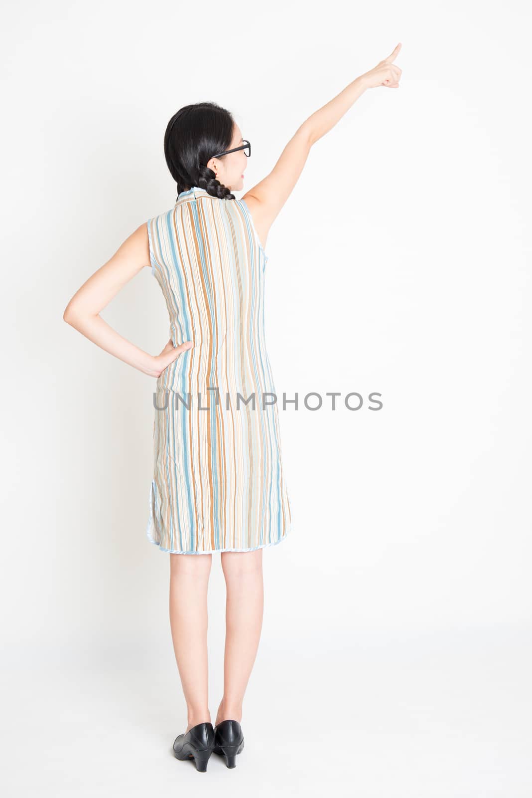 Rear view of young Asian girl in traditional qipao dress hand pointing away, full length standing on plain background.