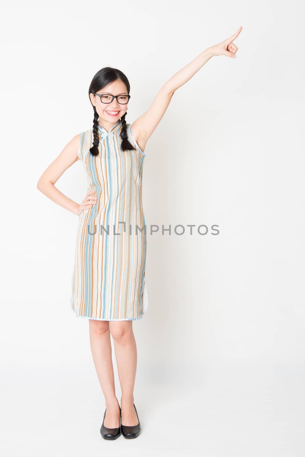 Portrait of young Asian girl in traditional qipao dress finger pointing away, full length standing on plain background.
