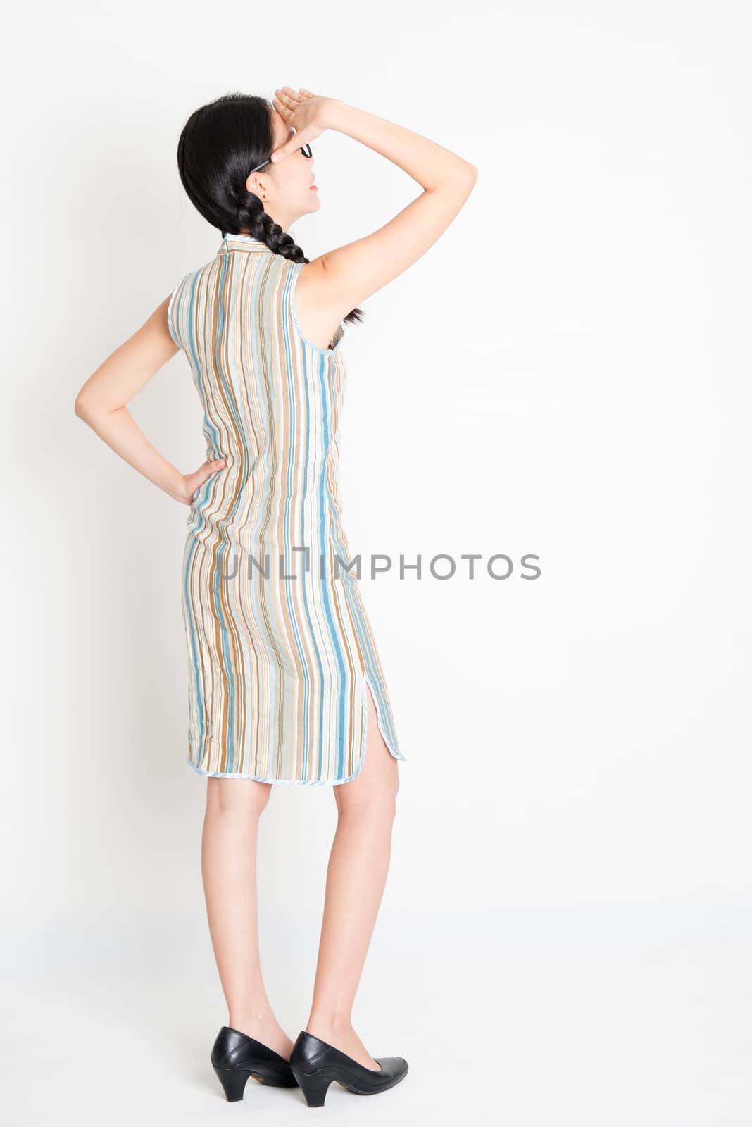 Rear view of young Asian woman in traditional qipao dress hand shielding, full length standing on plain background.