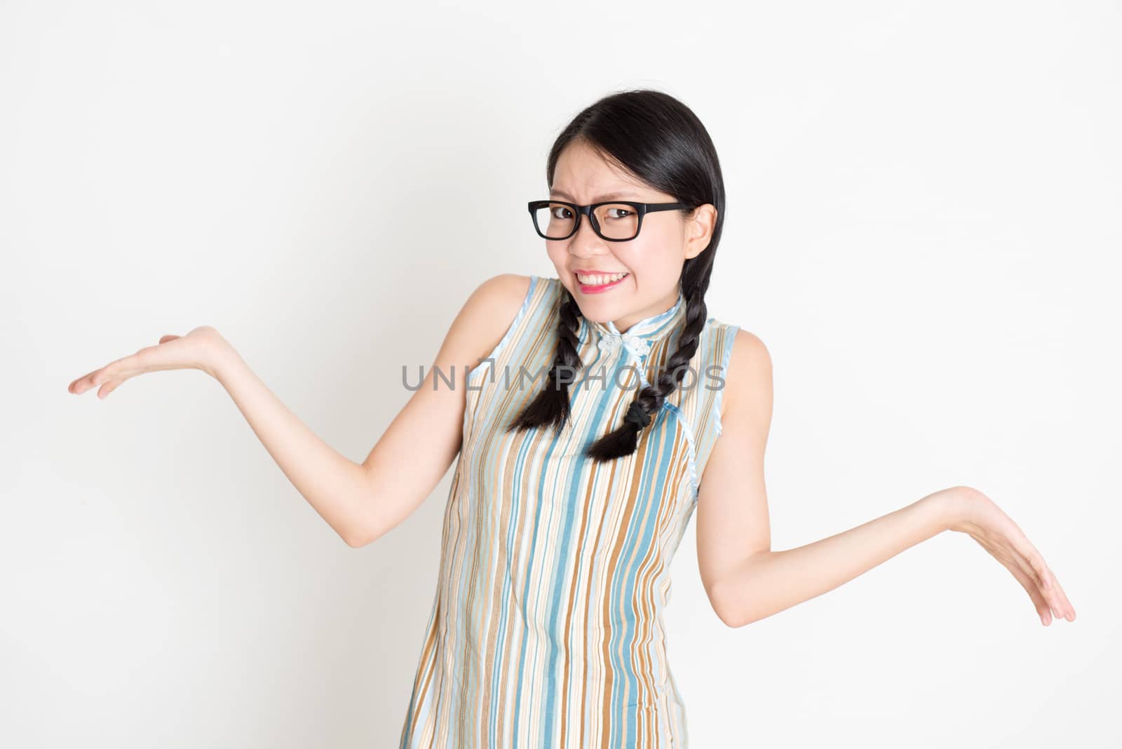 Portrait of young Asian girl in traditional qipao dress unsure and shrugging shoulder, standing on plain background.