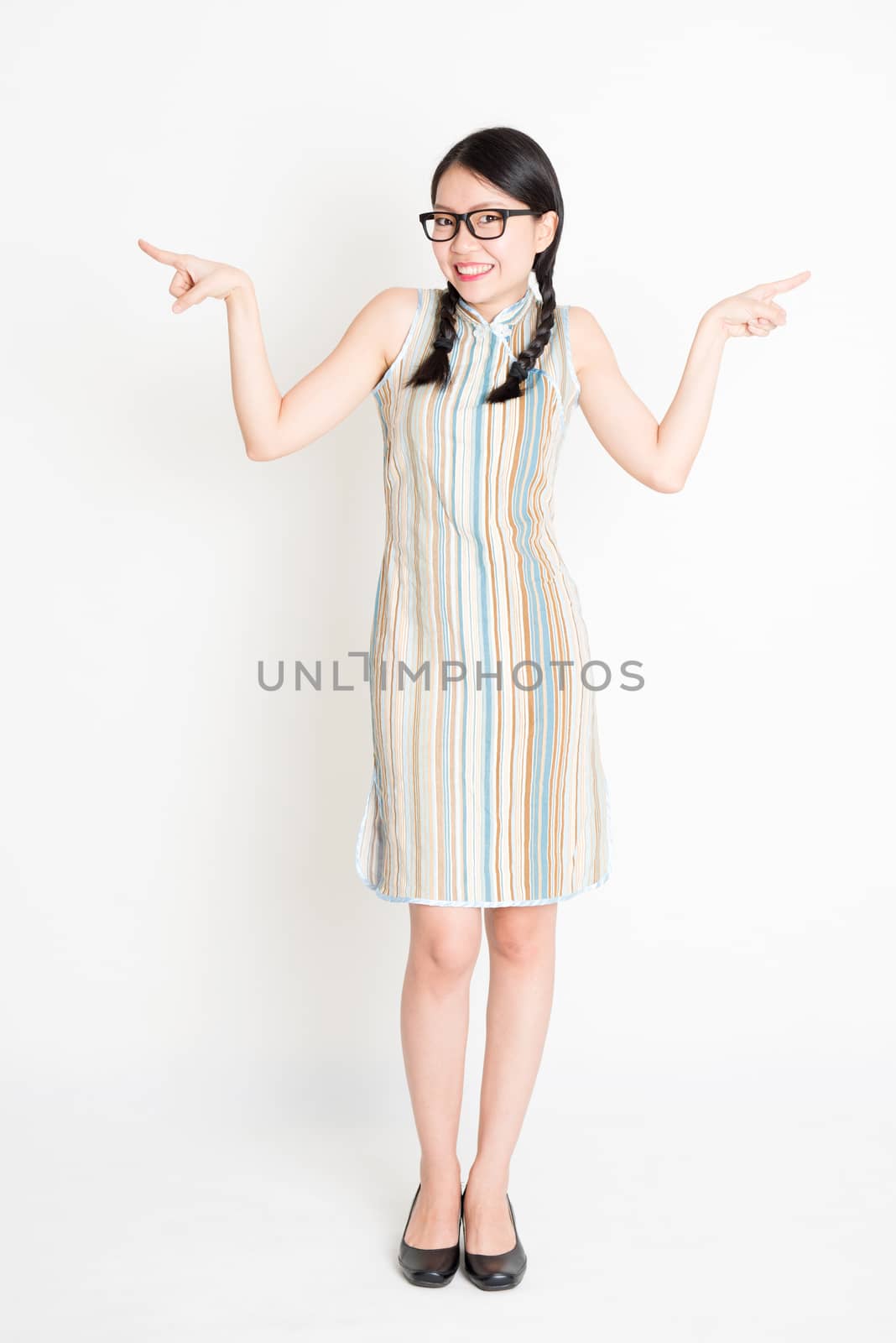 Portrait of young Asian woman in traditional qipao dress finger pointing at something, full length standing on plain background.