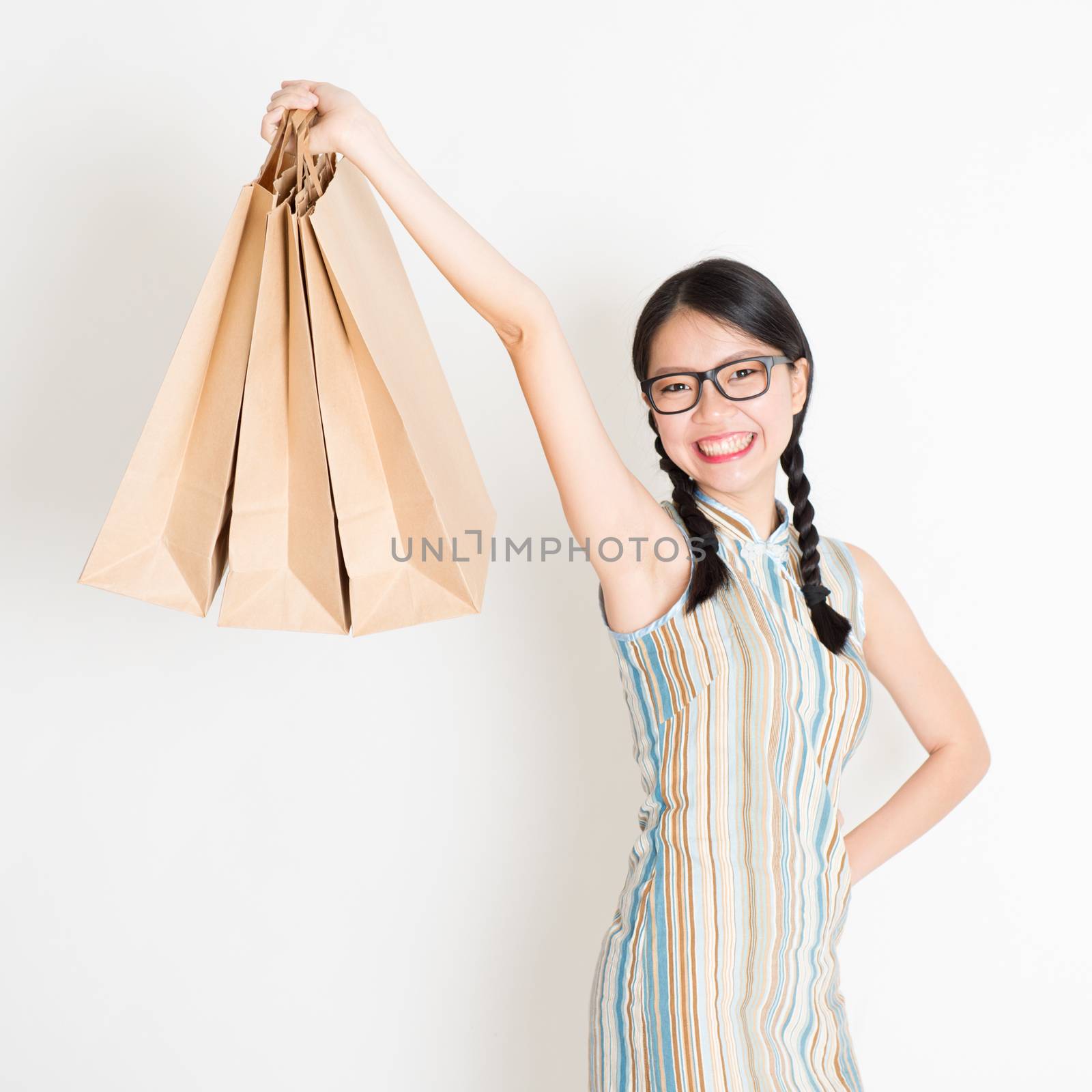 Portrait of young Asian female in traditional qipao dress shopping, hand holding paper bag, celebrating Chinese Lunar New Year or spring festival, standing on plain background.