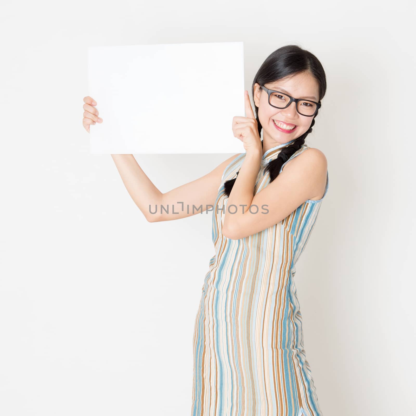 Portrait of young Asian girl in traditional qipao dress hand holding white blank paper card, celebrating Chinese Lunar New Year or spring festival, standing on plain background.