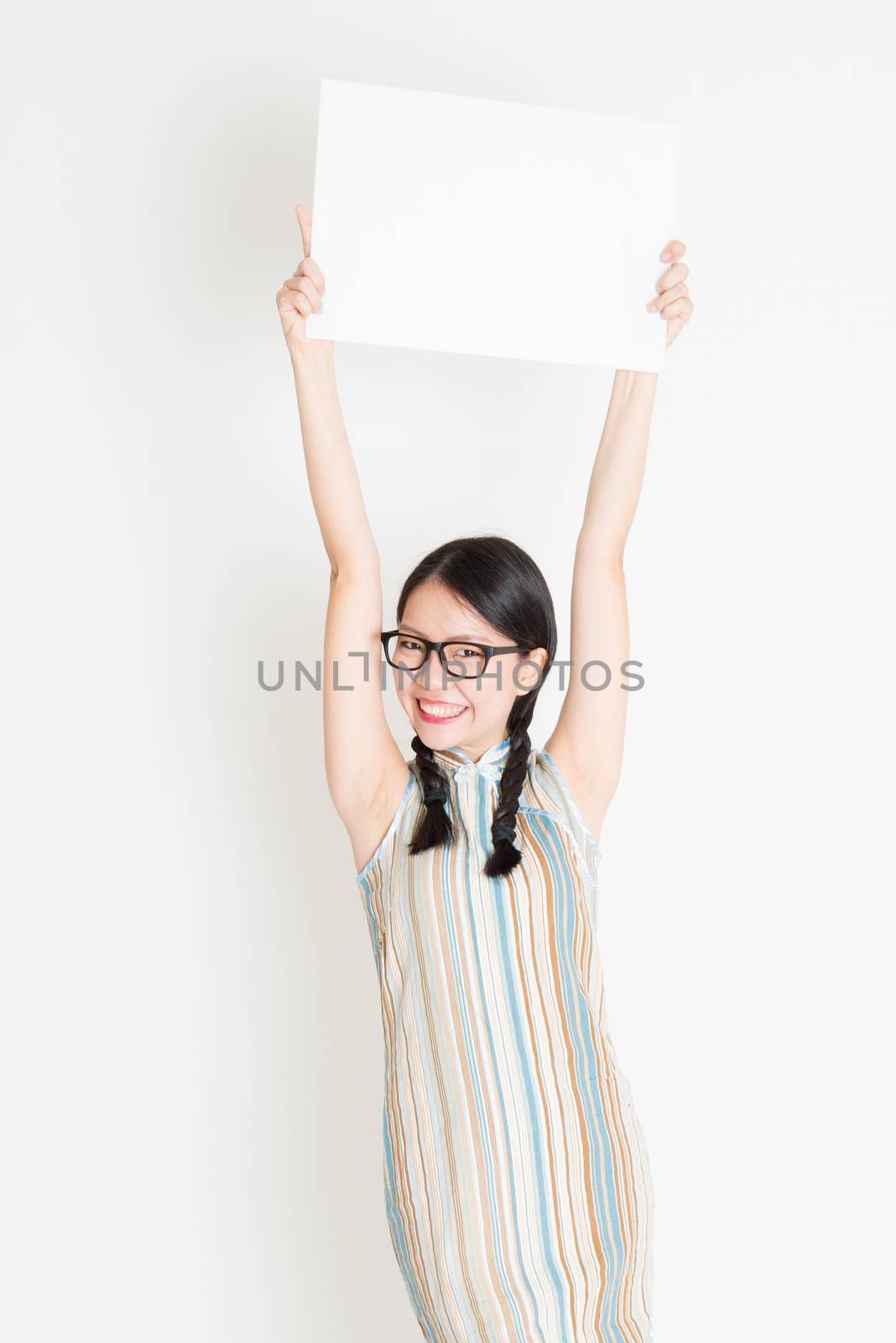 Portrait of young Asian woman in traditional qipao dress hand holding white blank paper card, celebrating Chinese Lunar New Year or spring festival, standing on plain background.