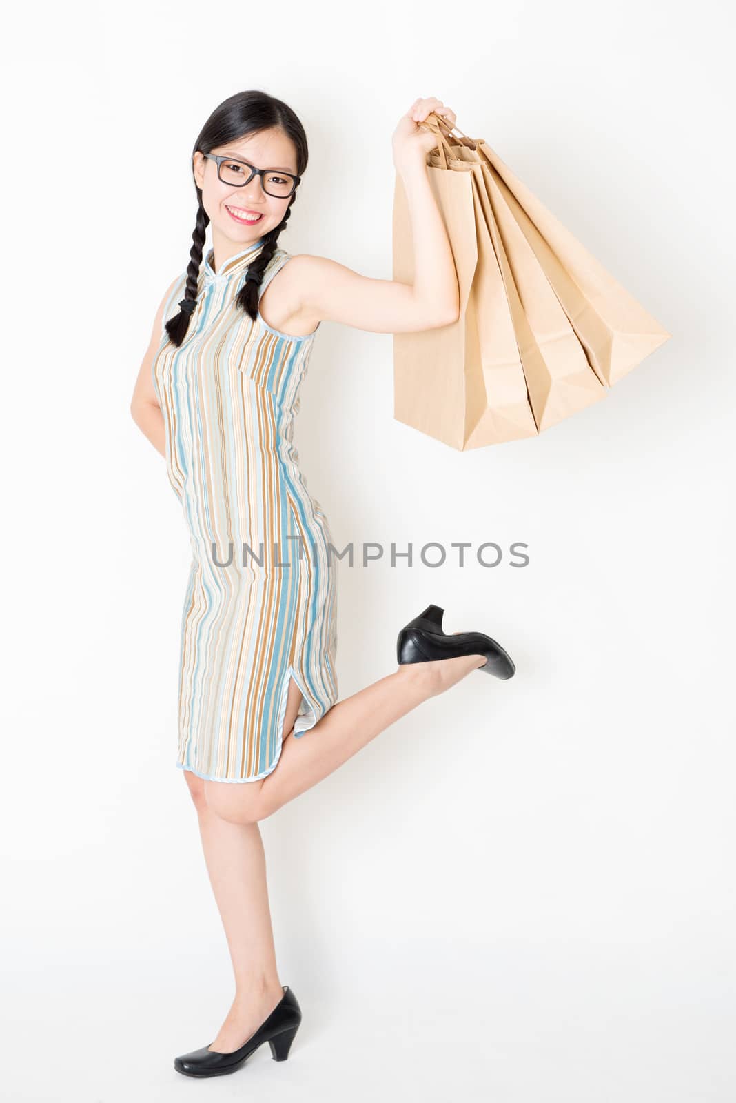 Portrait of young Asian girl in traditional cheongsam dress shopping, hand holding paper bag, celebrating Chinese Lunar New Year or spring festival, full length standing on plain background.