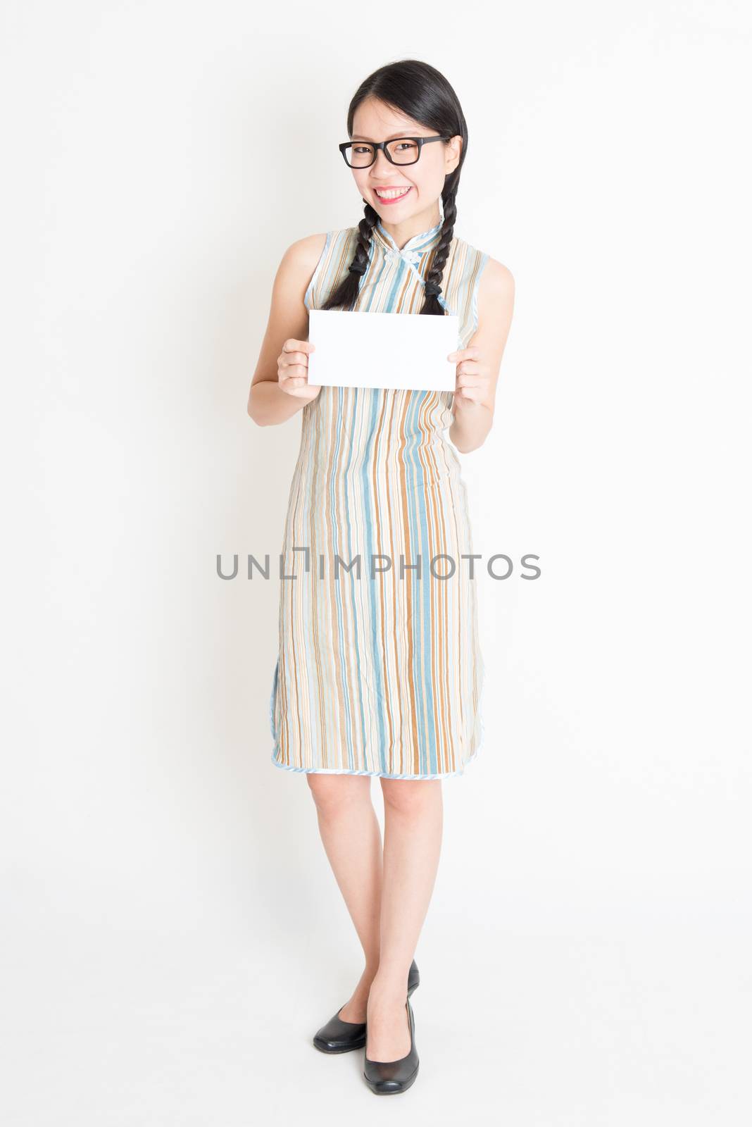 Portrait of young Asian female in traditional qipao dress hand holding white blank paper card, celebrating Chinese Lunar New Year or spring festival, full body standing on plain background.