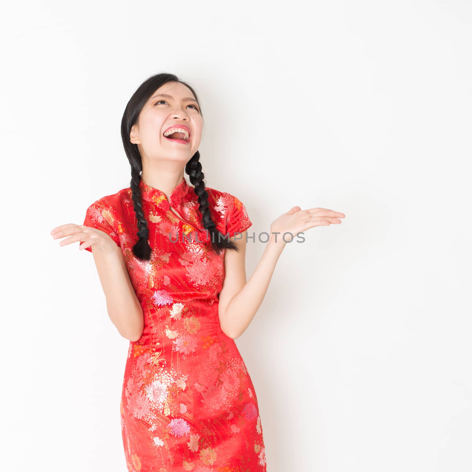 Portrait of surprise young Asian girl in traditional qipao dress with hand opened and looking up, celebrating Chinese Lunar New Year or spring festival, standing on plain background.