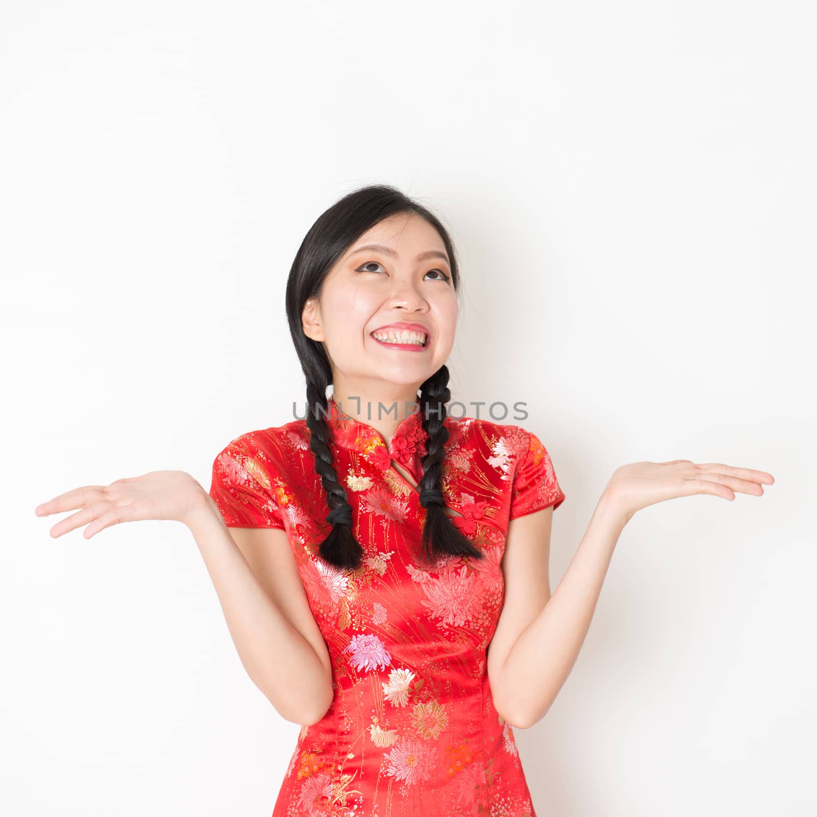Portrait of surprise young Asian woman in traditional qipao dress with hand opened and looking up, celebrating Chinese Lunar New Year or spring festival, standing on plain background.