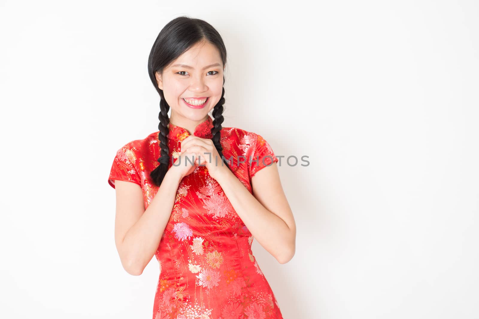 Portrait of young Asian girl in traditional qipao dress smiling and greeting, celebrating Chinese Lunar New Year or spring festival, standing on plain background.