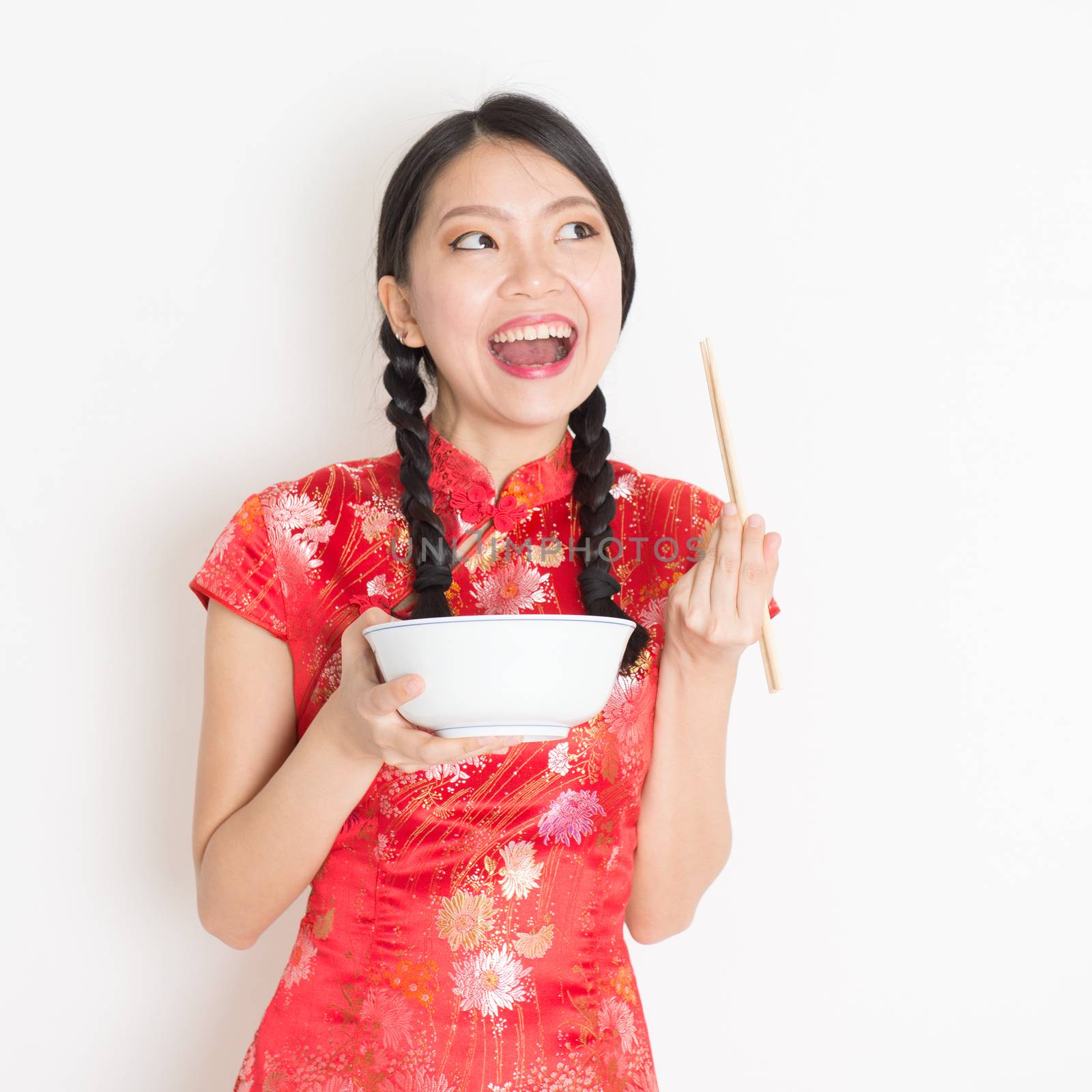 Portrait of young Asian woman in traditional cheongsam dress eating, hand holding bowl and chopsticks, celebrating Chinese Lunar New Year or spring festival, standing on plain background.