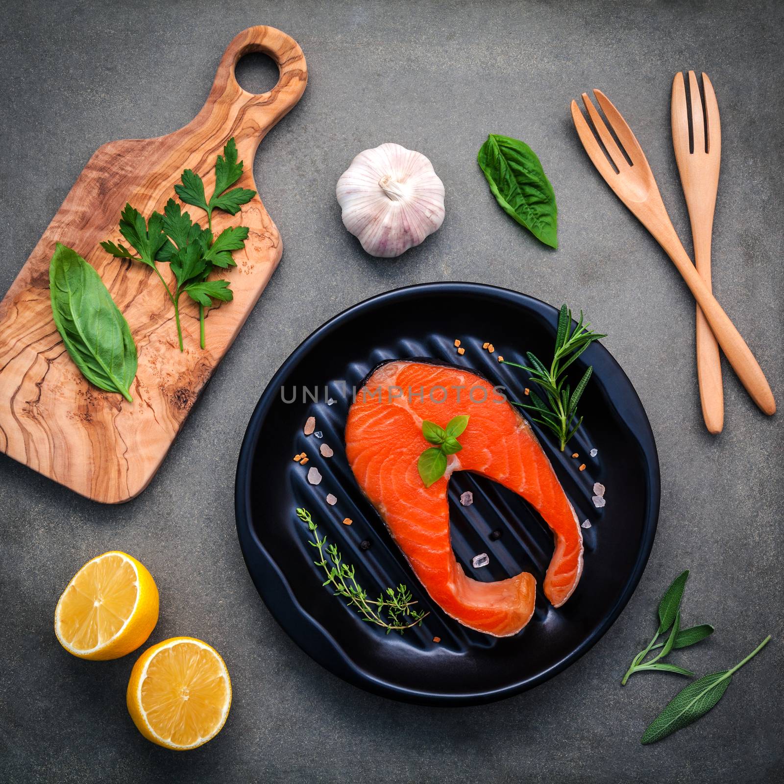Raw salmon fillet in the black plate with ingredients olive oil ,himalayan salt, and herbs sweet basil ,fennel ,sage ,rosemary ,garlic ,pepper and lemon  on dark concrete background .