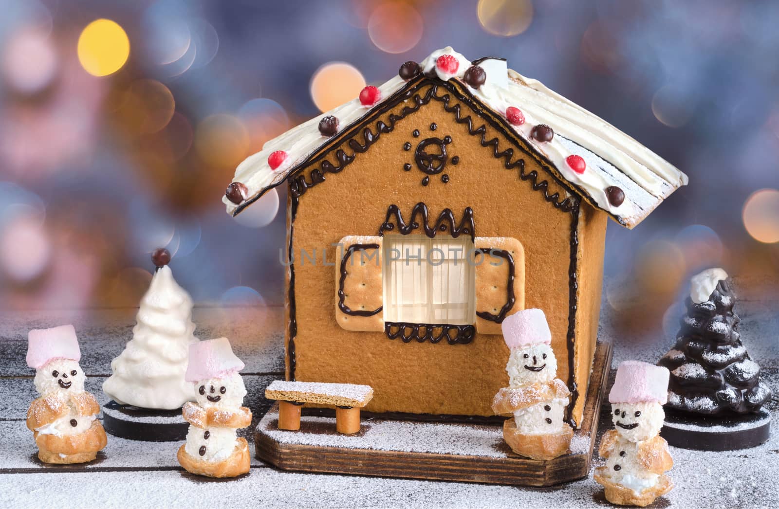 Christmas cake in the shape of the house, chocolate Christmas tree and sweet snowmen. On a wooden surface dusted with powdered sugar, colorful bokeh on the background