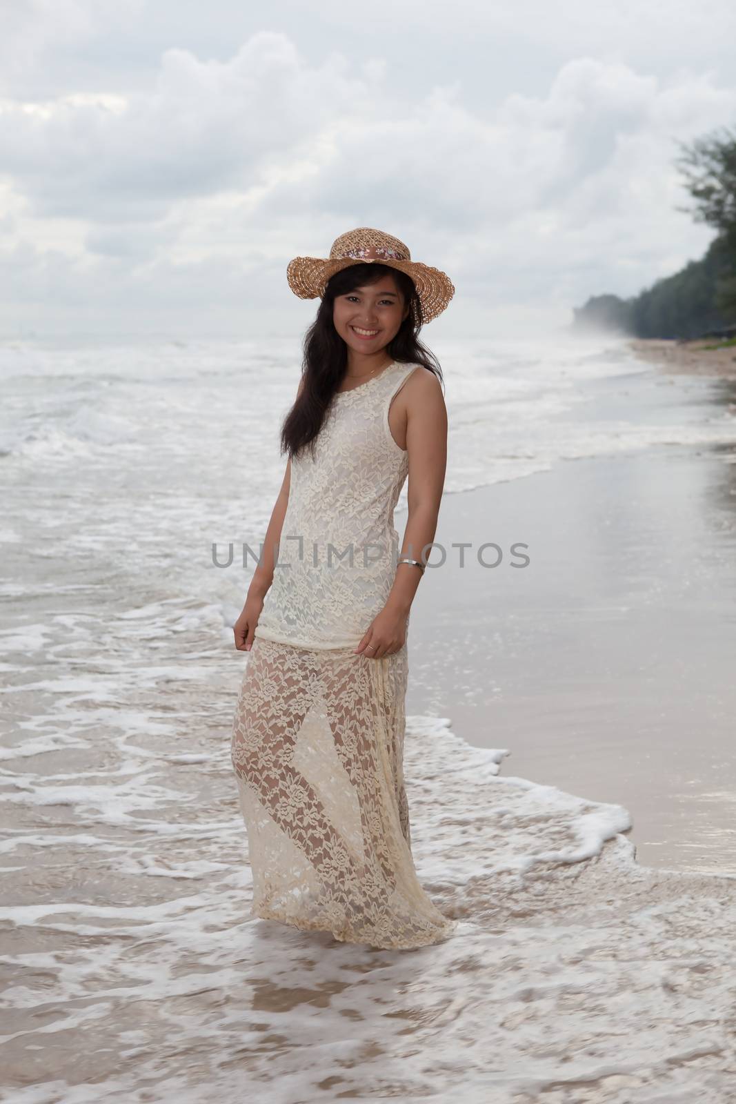 portrait of smiling face of young asian woman with long dress and fashion wide straw hat standing on sea beach 