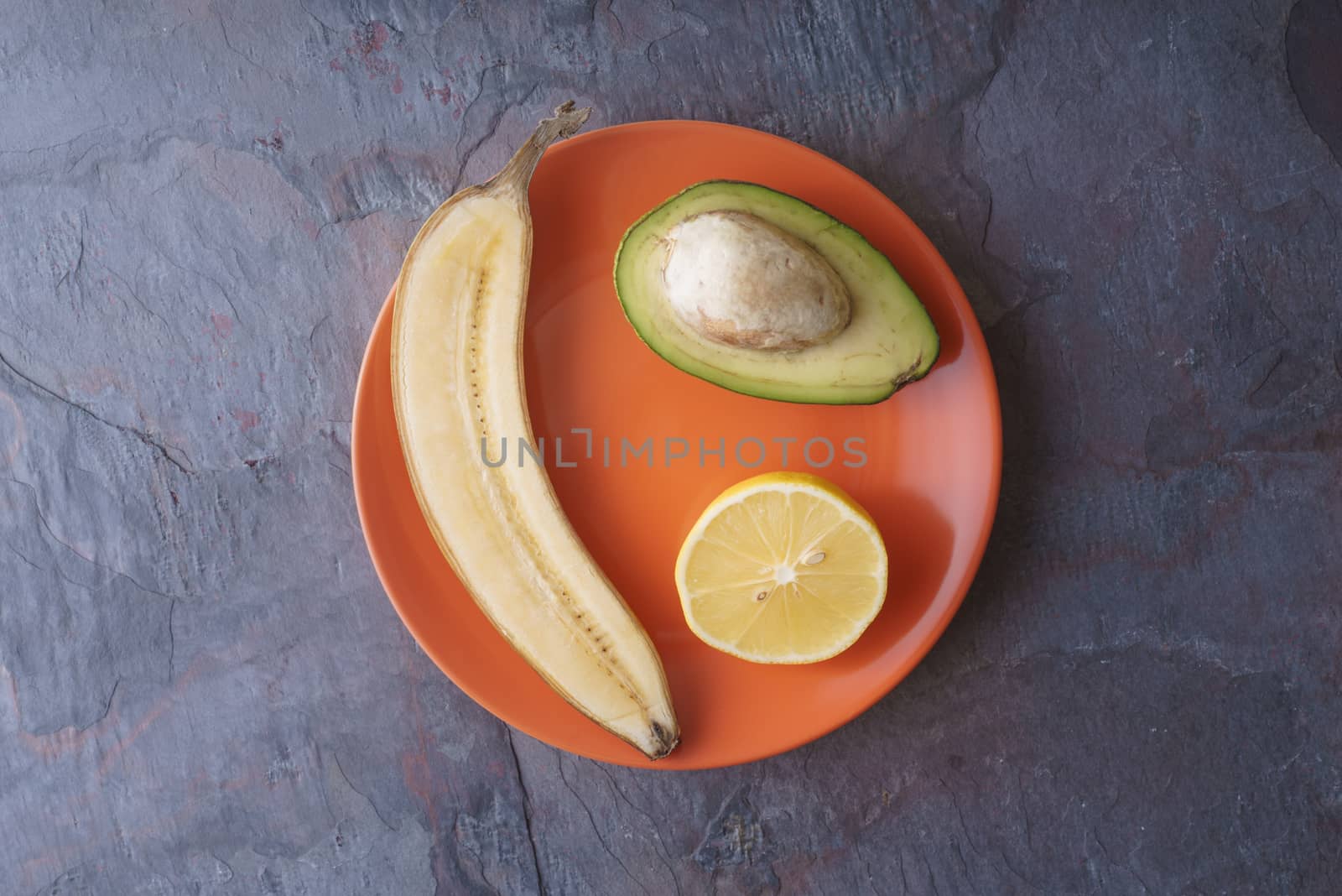 Avocado, lemon, banana halves lie on an orange plate on a slate horizontal