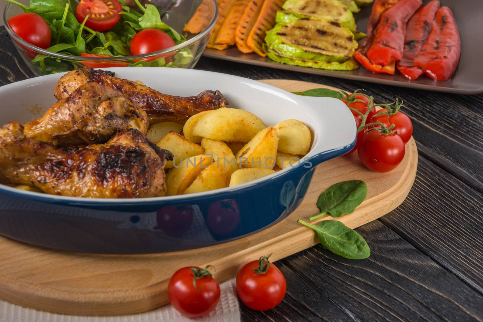 Tasty and nutritious meal with baked chicken drumsticks and potato in ceramic dish,  grilled vegetables, salad and tomatoes on the background of dark table