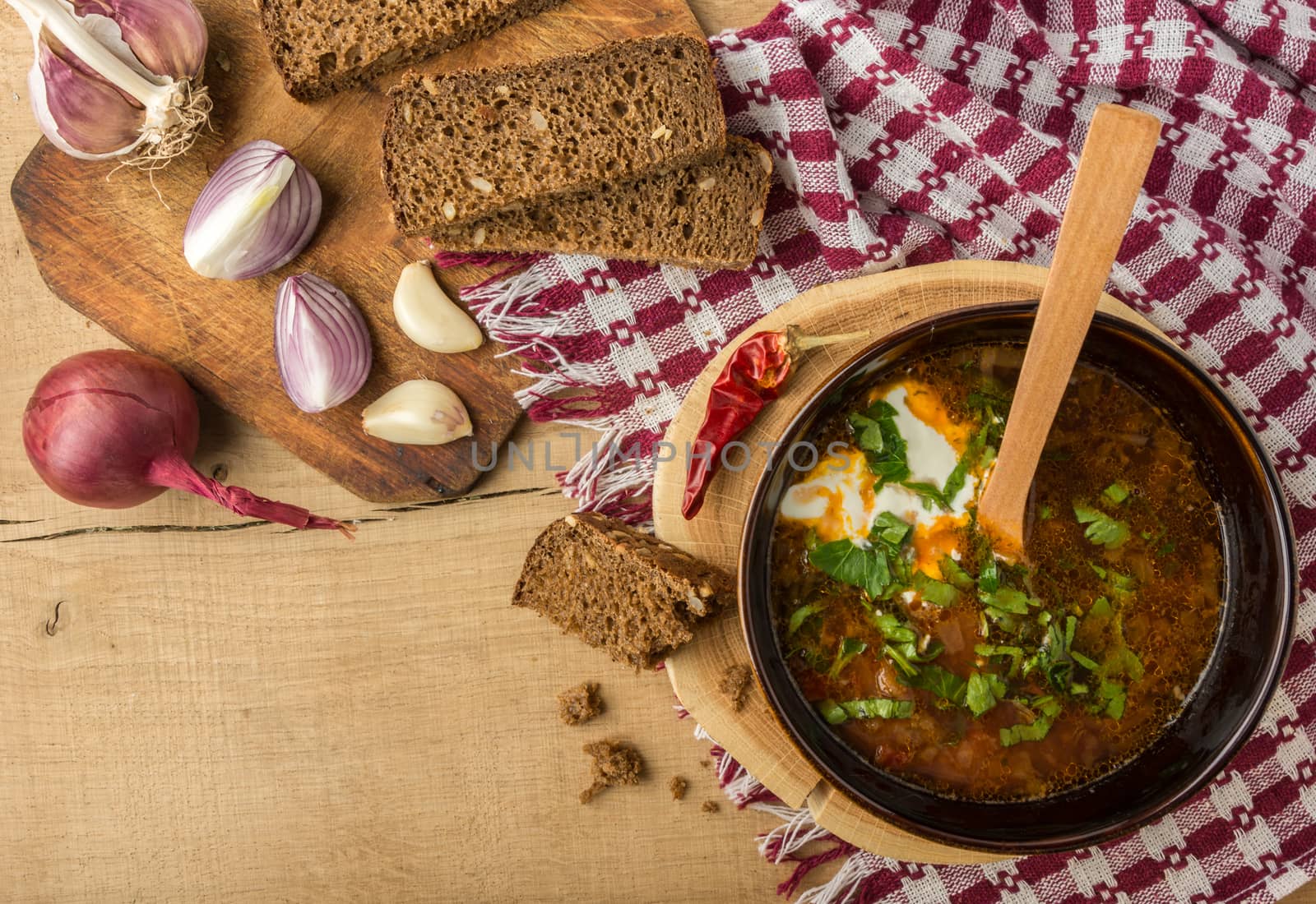 Tasty and nutritious soup with meat, potatoes, cabbage, tomatoes, beans, sour cream, parsley, onion, garlic, dark bread in a clay plate on a wooden tray and wooden table. Top view