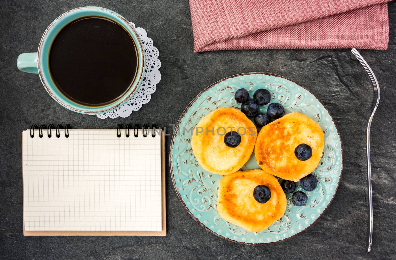 cheese pancake with blueberries, powdered sugar, notebook and black coffee on a dark background top view