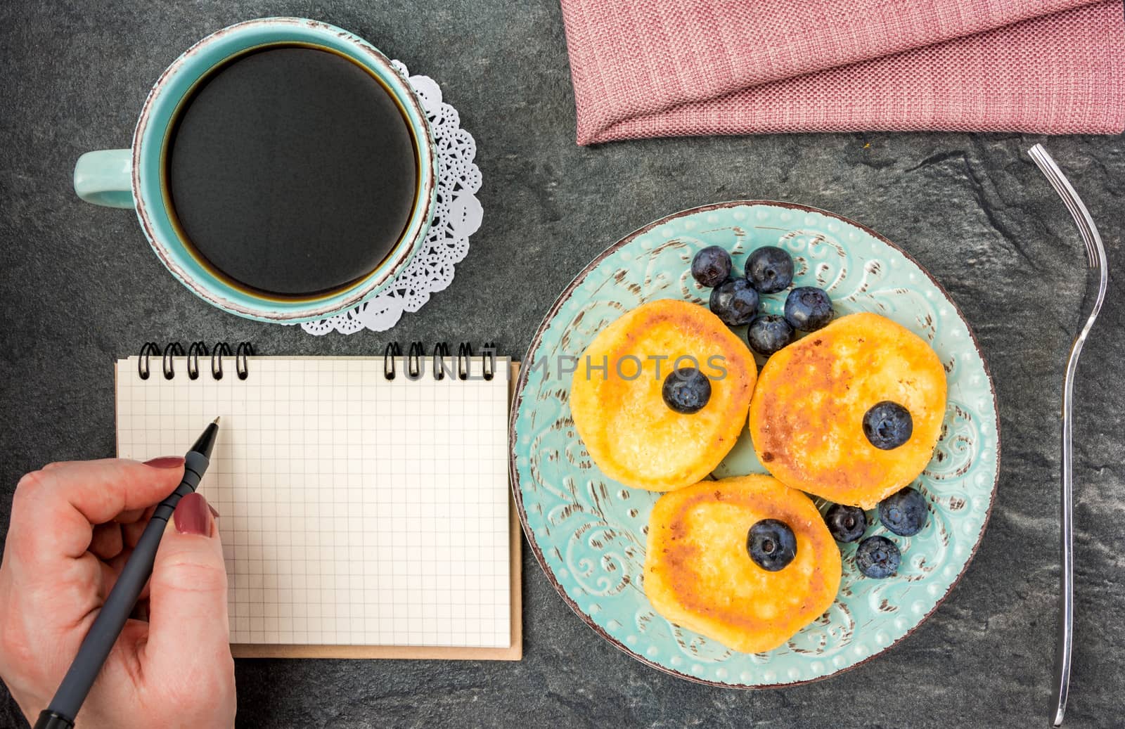 Woman having breakfast with cheese pancake with blueberries, powdered sugar, black coffee on a dark wooden table and writing in a notebook . Top view.