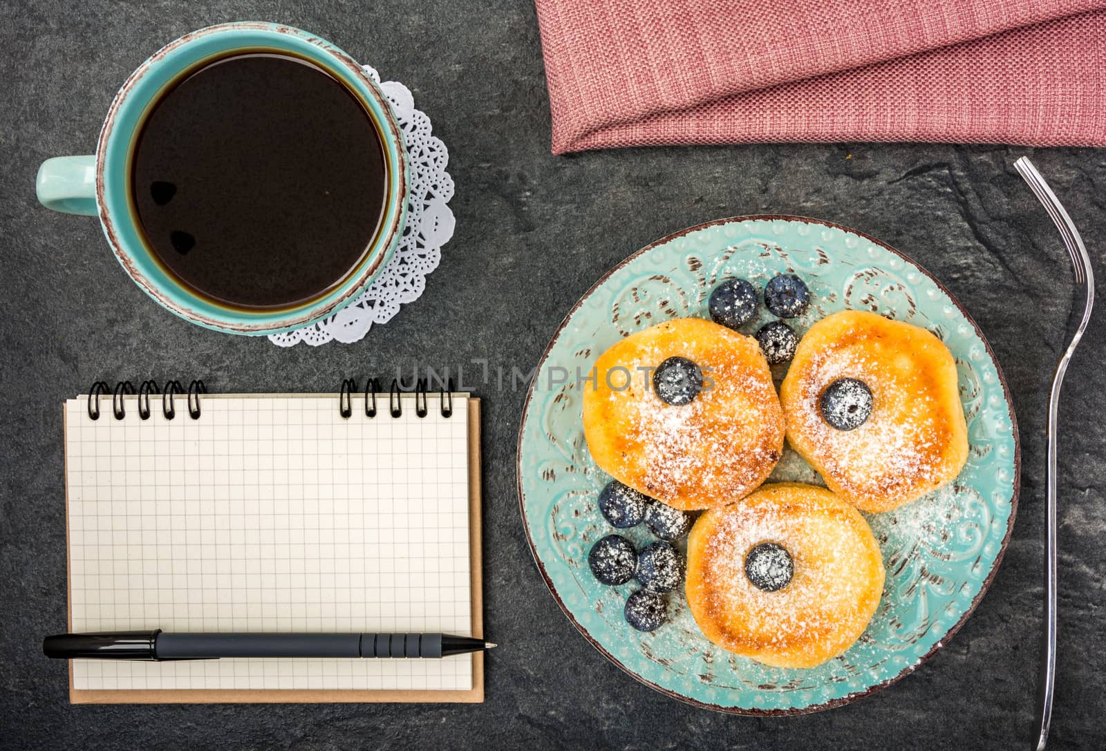 cheese pancake with blueberries, powdered sugar, notebook, pen and black coffee on a dark background top view