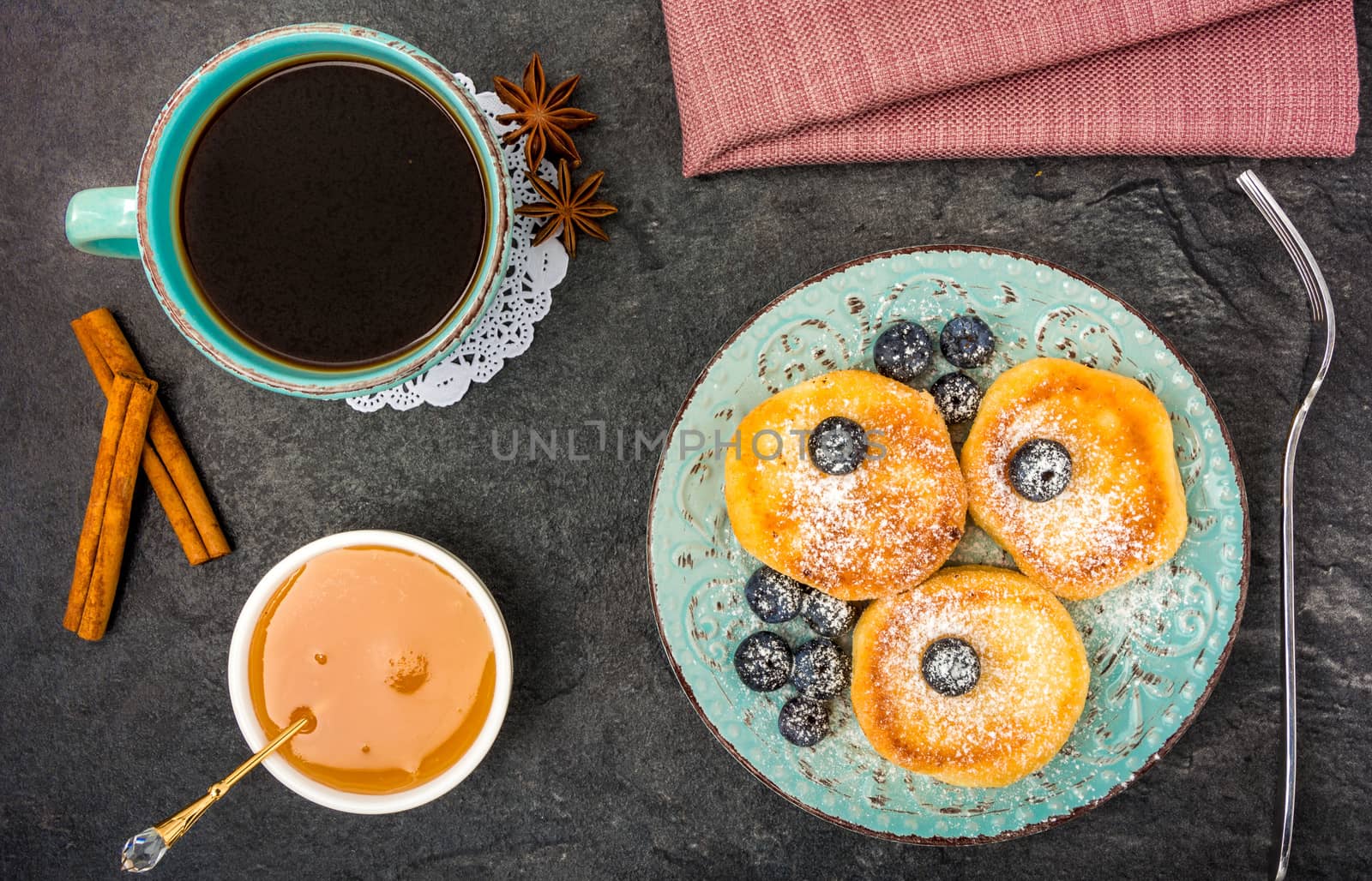 cheese pancake with blueberries, powdered sugar, honey and black coffee on a dark background top view