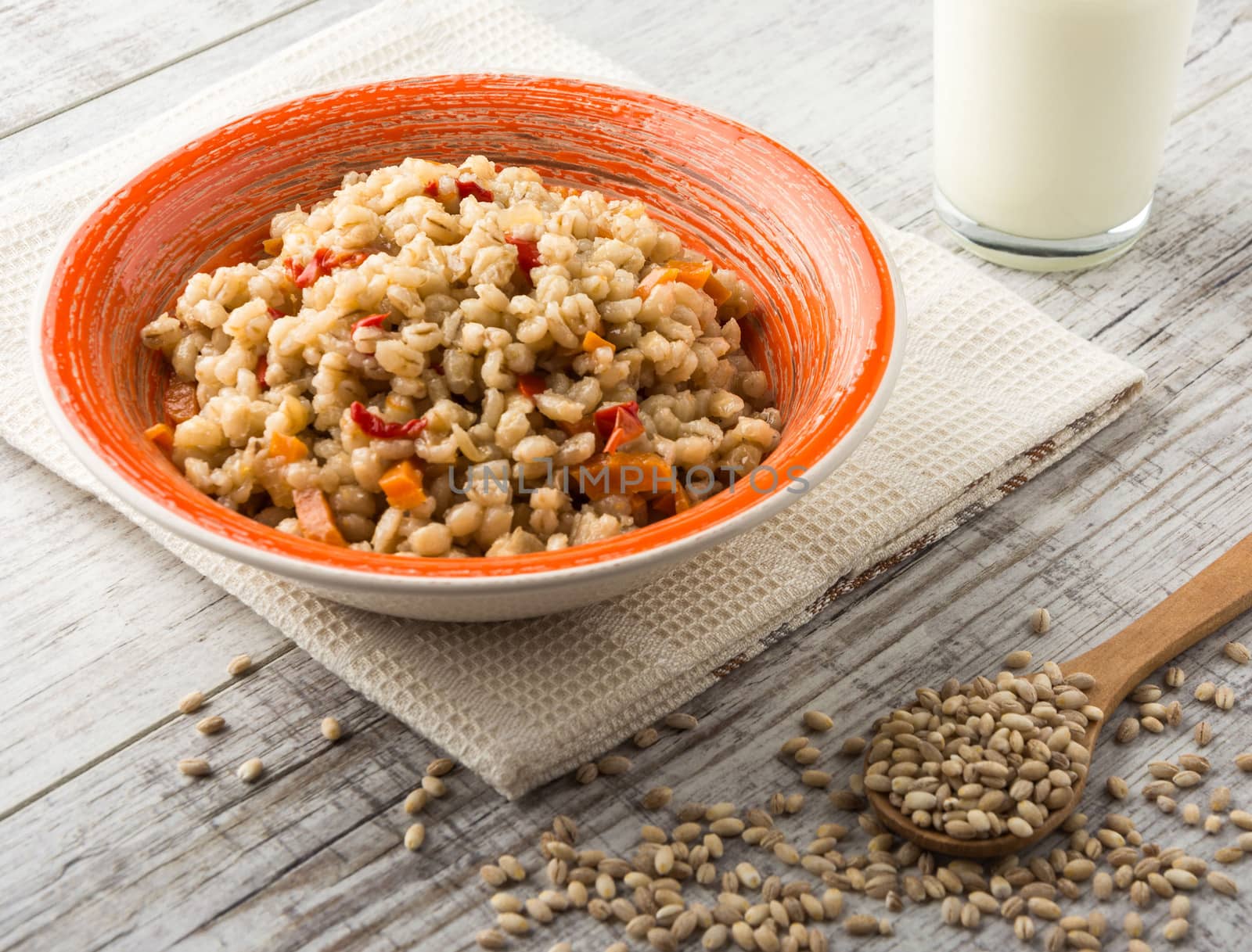 Pearl barley porridge with vegetables in an orange ceramic plate with a kitchen towel, pearl barley in a wooden spoon and glass of milk on a white wooden table