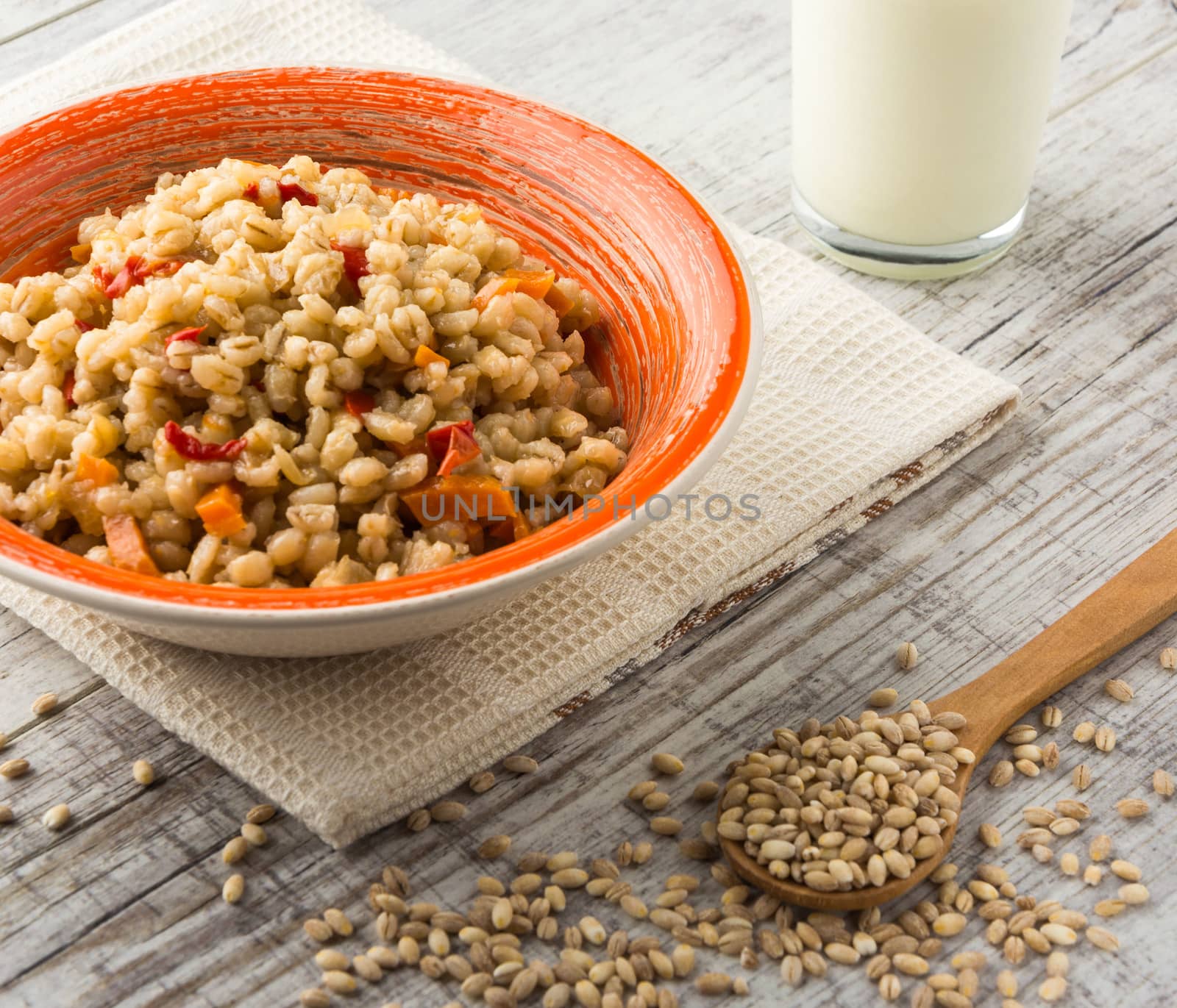 Pearl barley porridge with vegetables in an orange ceramic plate with a kitchen towel, pearl barley in a wooden spoon and glass of milk on a white wooden table