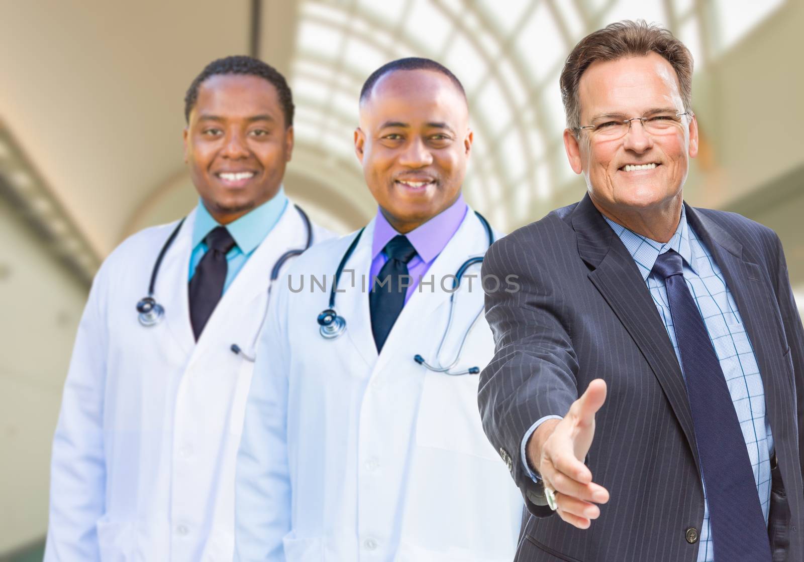 Two Mixed Race Doctors Behind Businessman Reaching for a Hand Shake Inside Hospital.