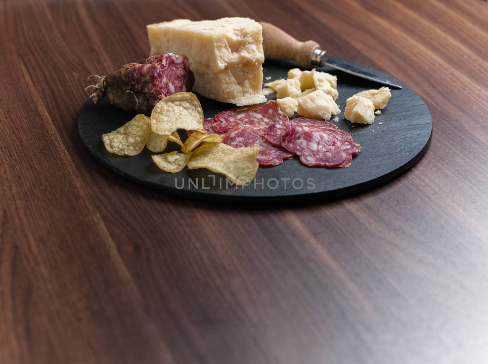 A dark stone tray on a wooden kitchen table filled with parmesan cheese, salami and potato chips.