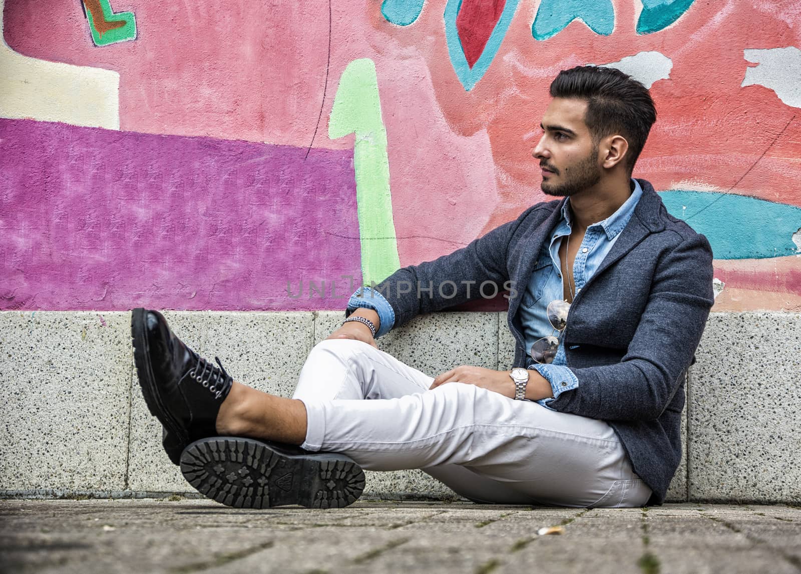 Attractive young man sitting against colorful graffiti wall, looking away