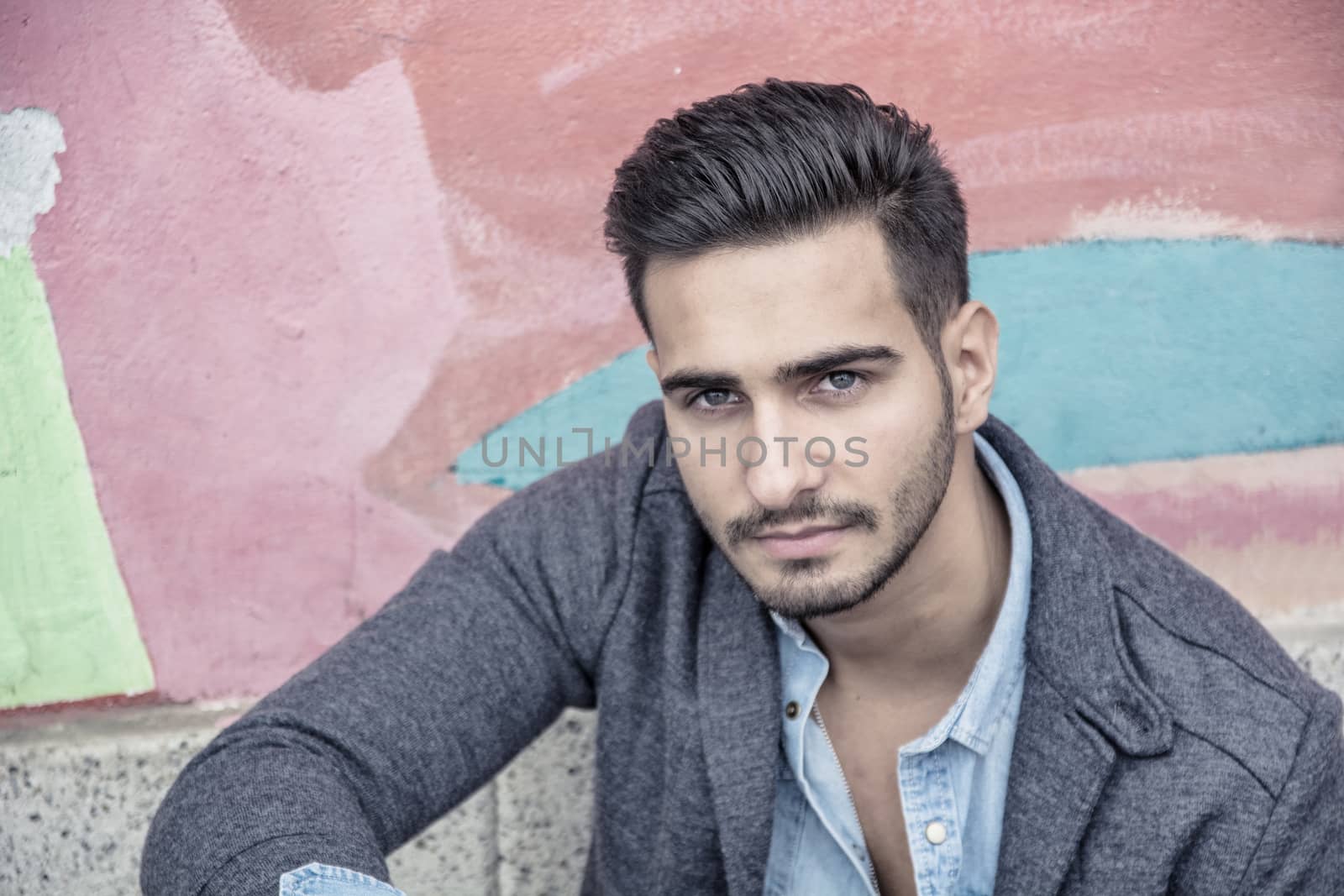 Attractive young man sitting against colorful graffiti wall, looking at camera