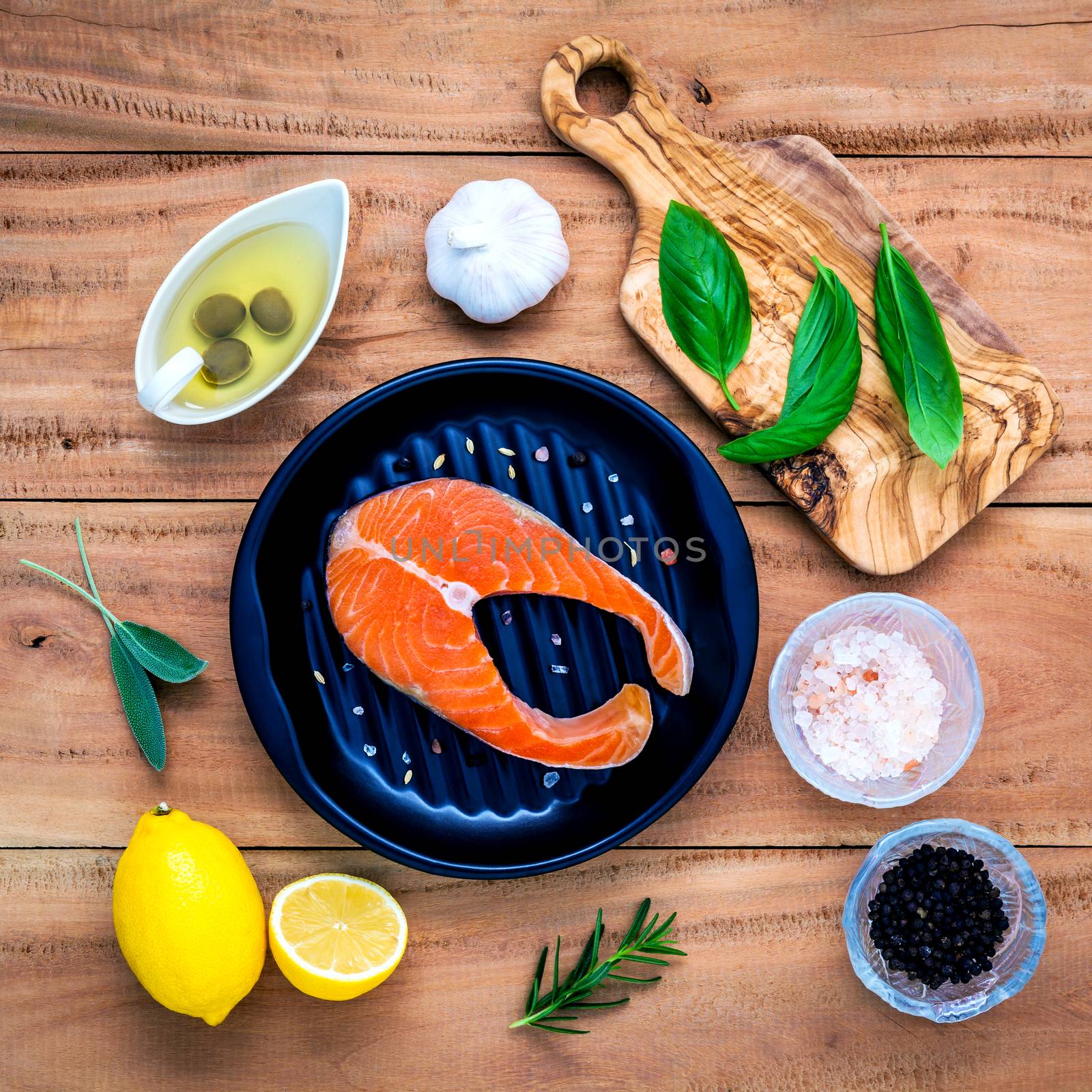 Raw salmon fillet in the black plate with ingredients olive oil ,himalayan salt, and herbs sweet basil ,fennel ,sage ,rosemary ,garlic ,pepper and lemon  on wooden background .