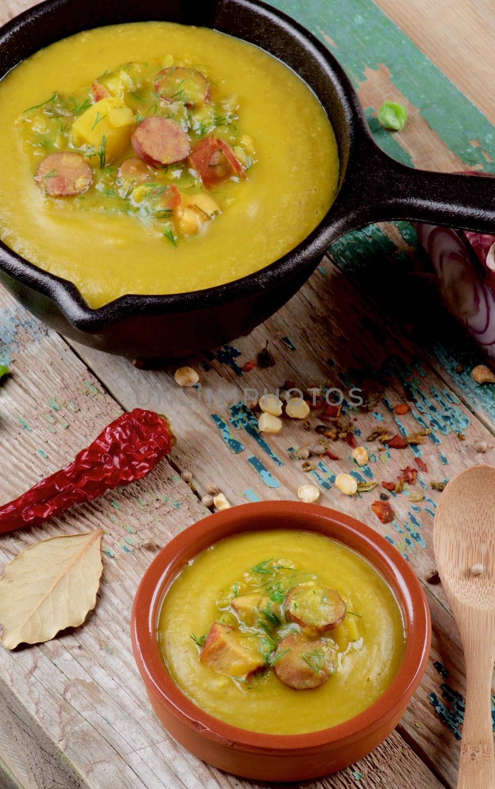 Pea Soup with Smoked Sausages in Black Iron Cast and Clay Bowl with Raw Pea, Spices and Wooden Spoon closeup on Cracked Wooden background