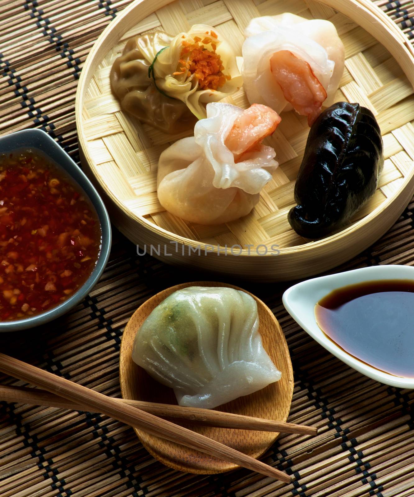Assorted Dim Sum in Bamboo Steamed Bowl and Hagao with Shrimp on Wooden Plate with Red Chili and Soy Sauces and Chopsticks closeup on Straw Mat background